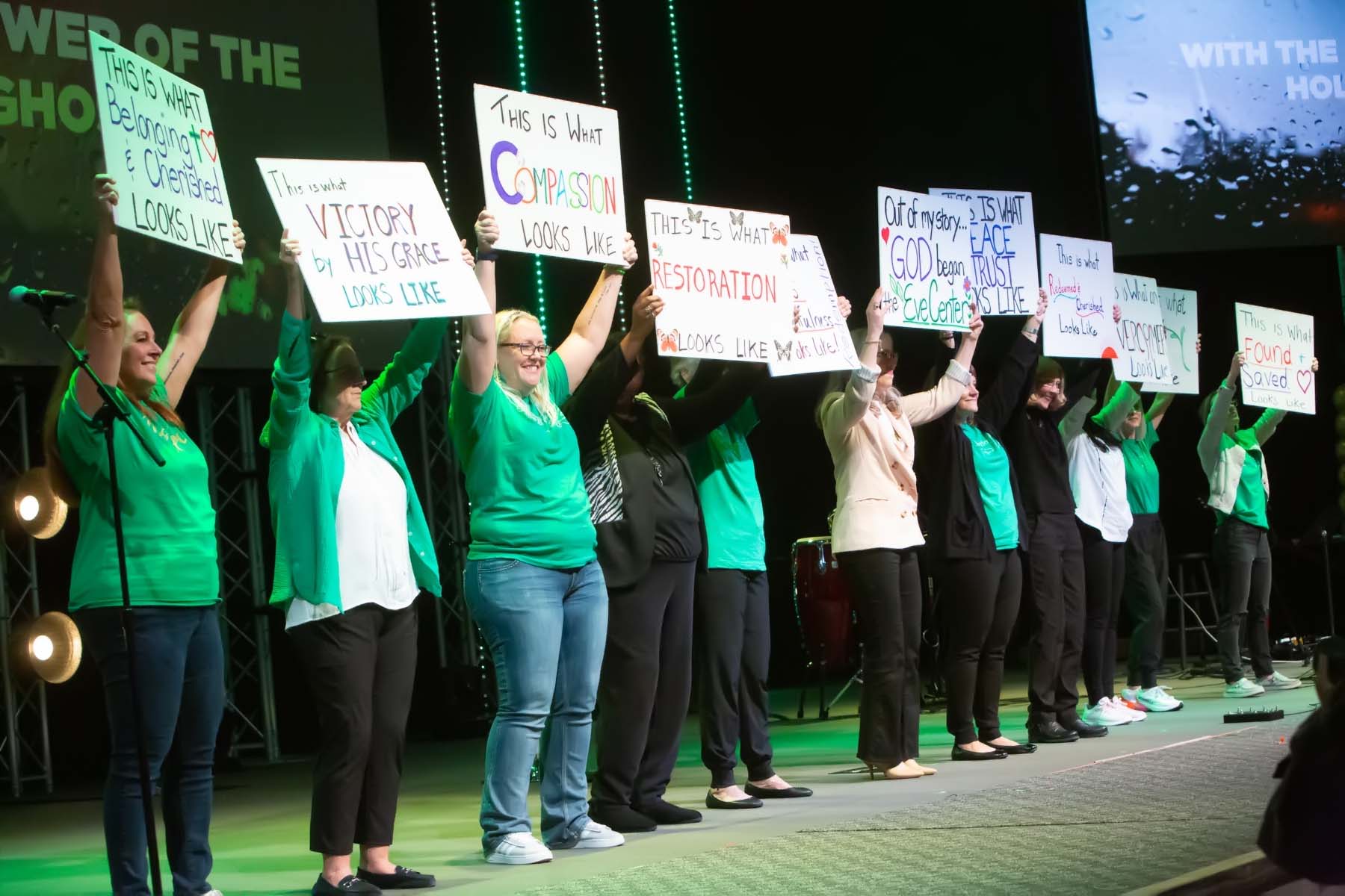 Group of people on stage holding signs during a protest or rally, with a green and black background and stage lighting.