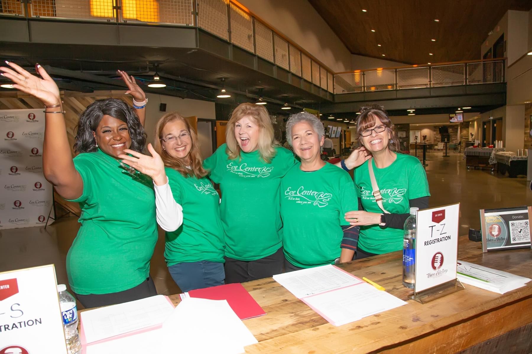 Five women in green shirts standing behind a table, smiling and waving indoors.