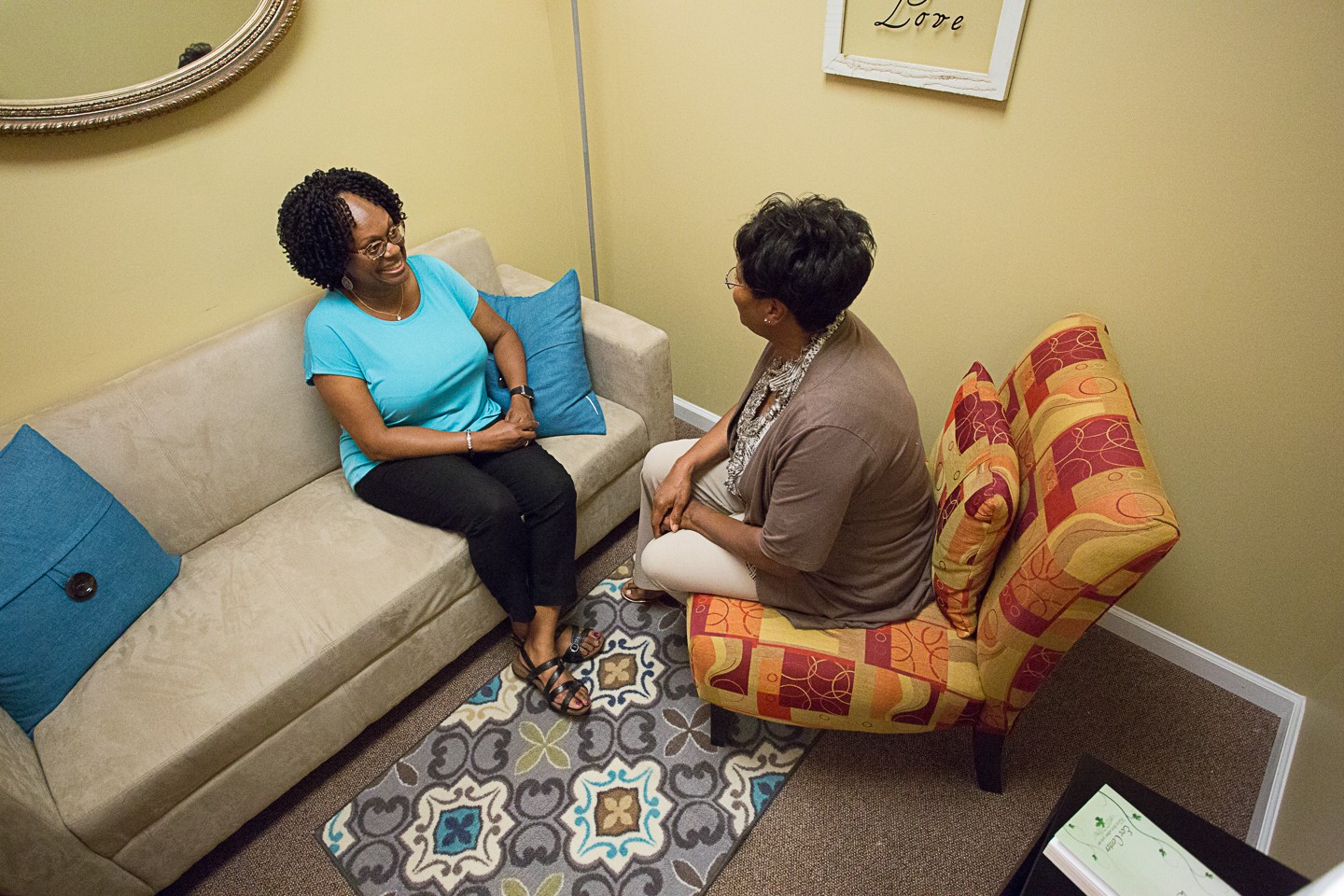 Two women sit and converse in a small room with yellow walls, a patterned carpet, and a beige sofa with blue pillows.