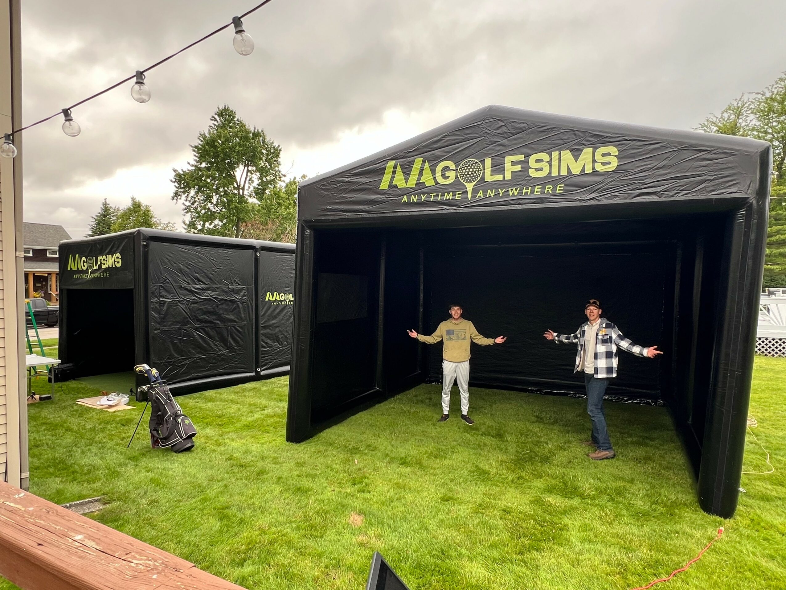 Two people stand inside a black inflatable structure with yellow text, outdoors on grass, with trees and a cloudy sky in background.