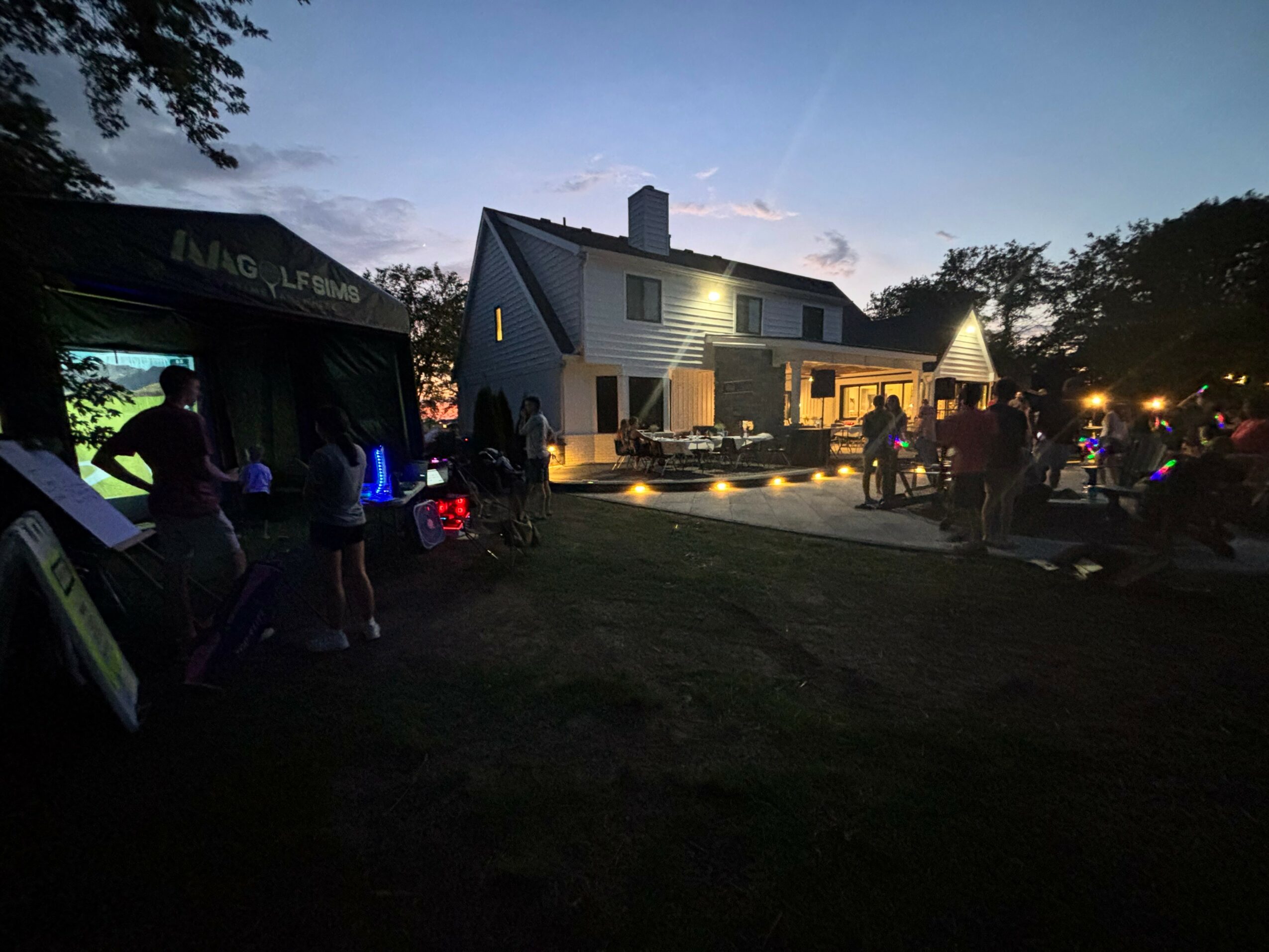 House with lit porch and people gathered outside during dusk, trees and sky in background.