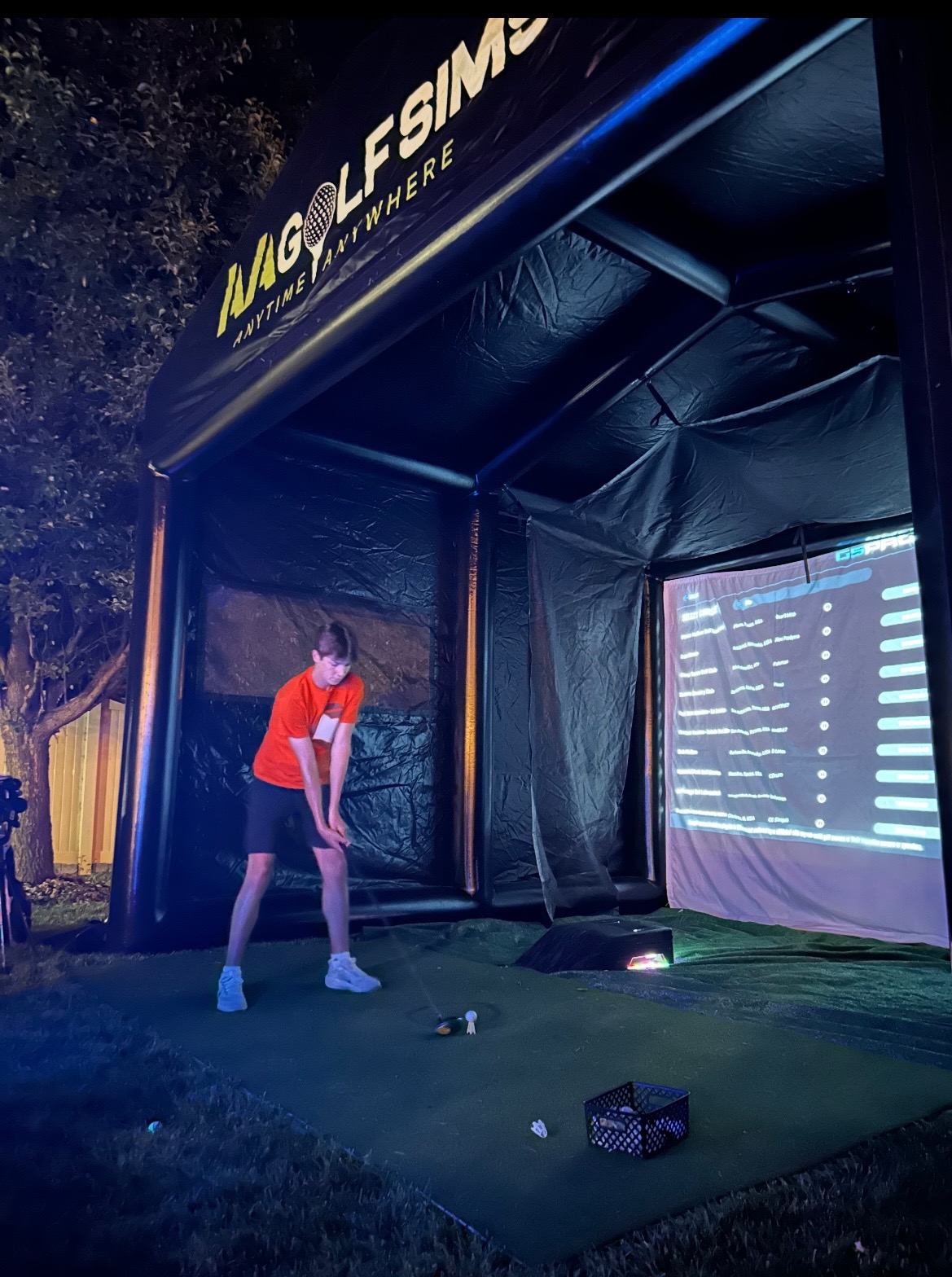 Person in red shirt and shorts standing inside a black indoor golf simulation tent with a large screen displaying data.