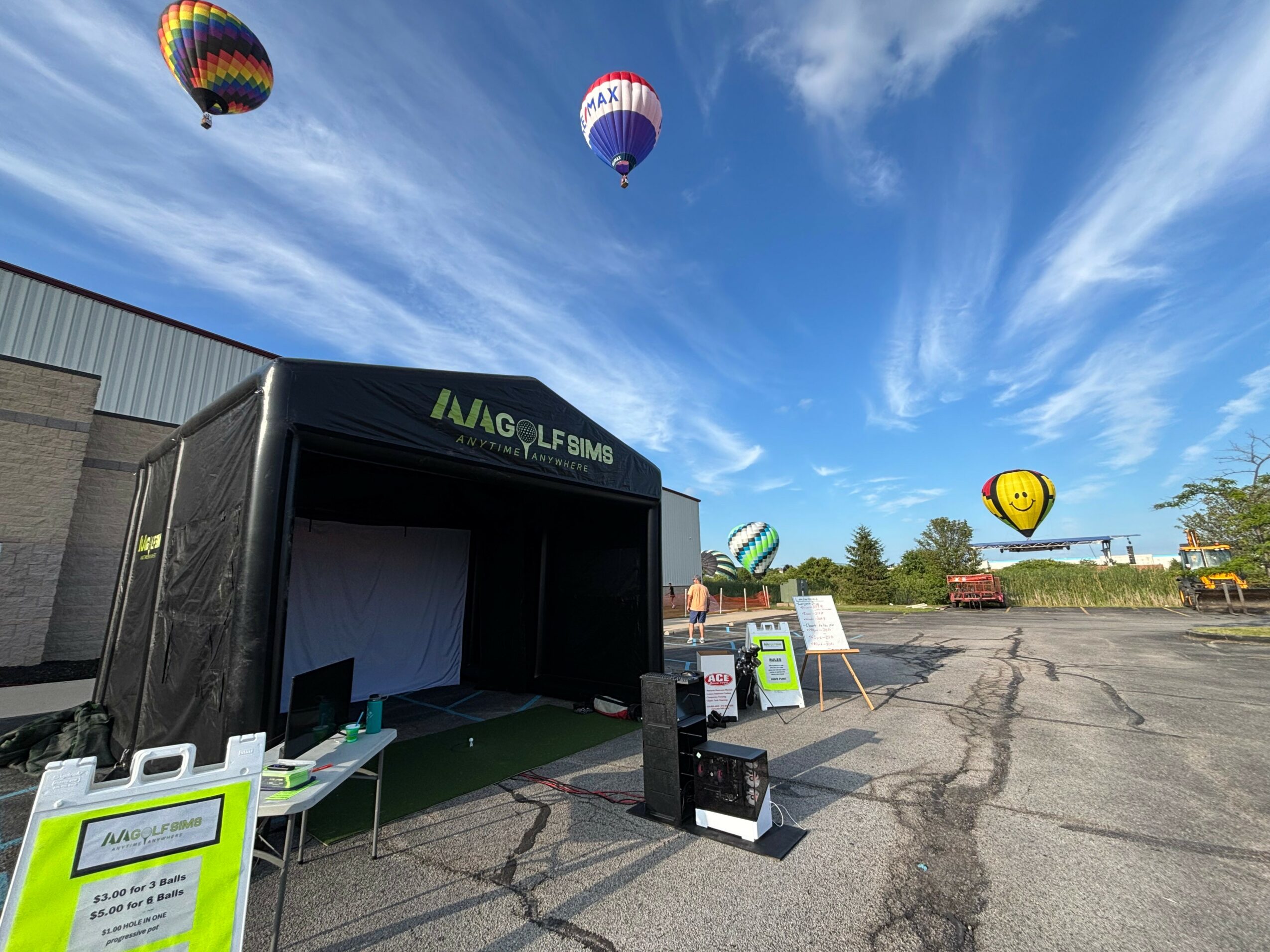 Hot air balloons in the sky above an outdoor event area with a black tent and signs.
