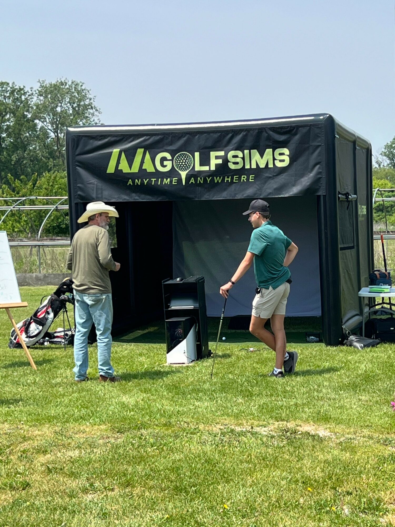 Two people stand outside a black tent with a sign reading 'MAGOLF SIMS' on a grassy area, one holding a golf club.