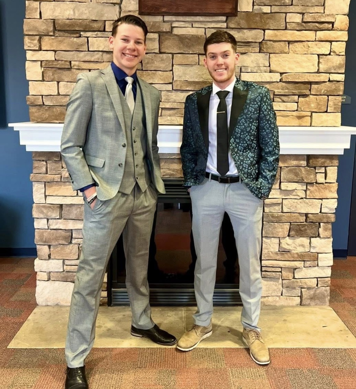 Two men in suits standing in front of a brick fireplace, smiling, with hands in pockets.