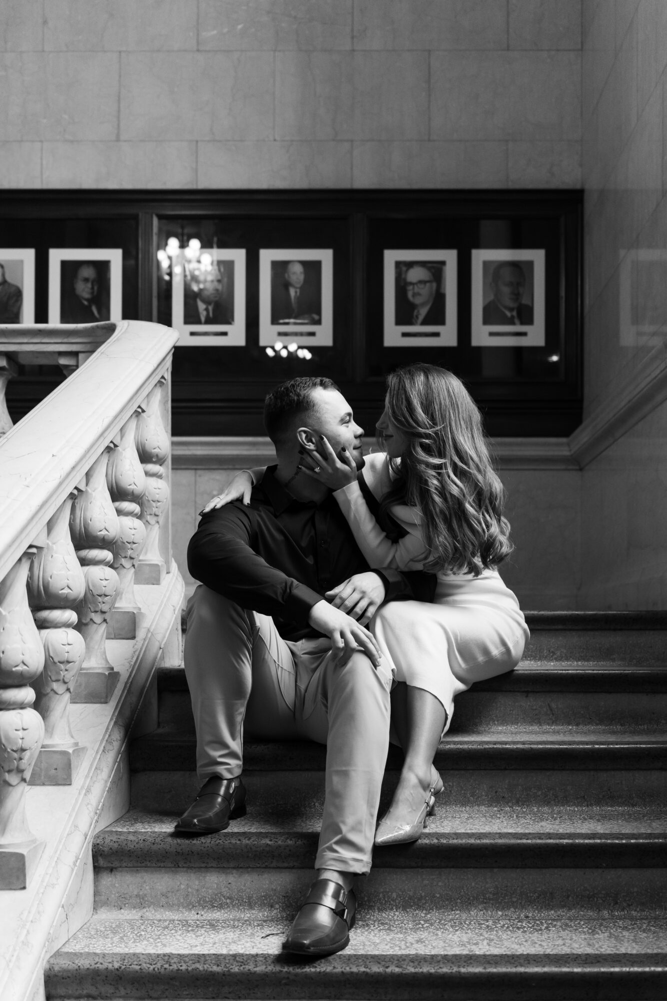 A couple sits on stairs in a gallery, looking at each other, with framed photos on the wall behind them.