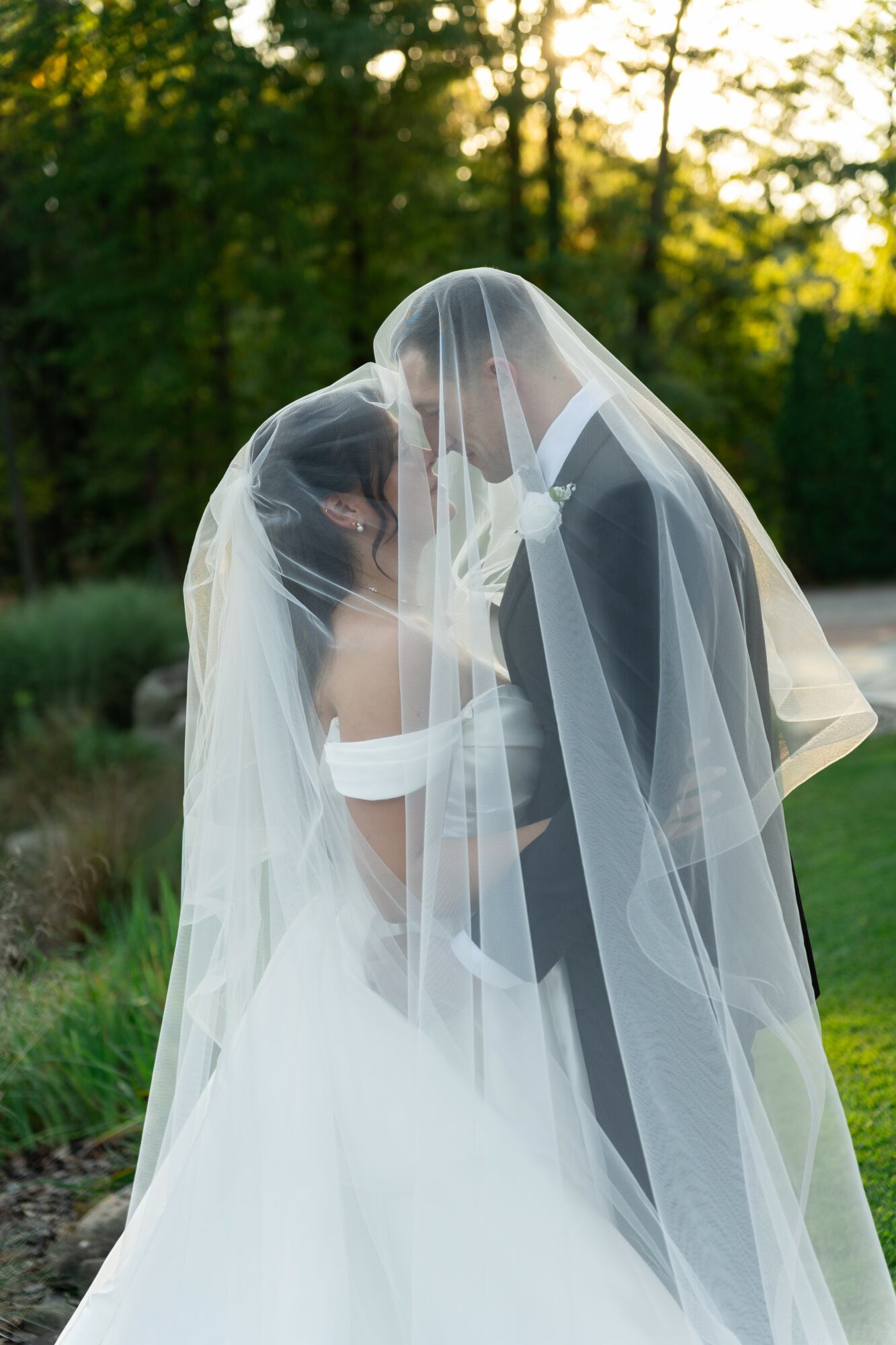 Bride and groom face each other under a veil outdoors with trees and sunlight in the background.