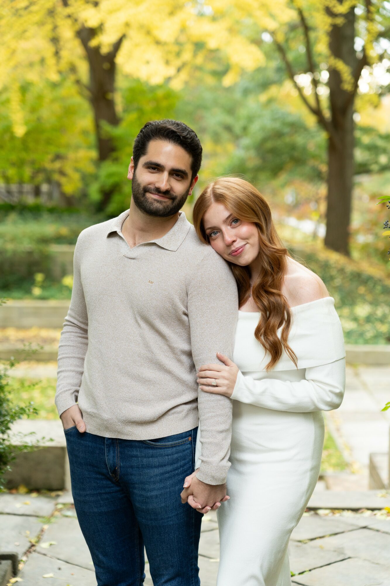 A smiling man and woman stand close together outdoors in autumn, with trees and a pond in the background.