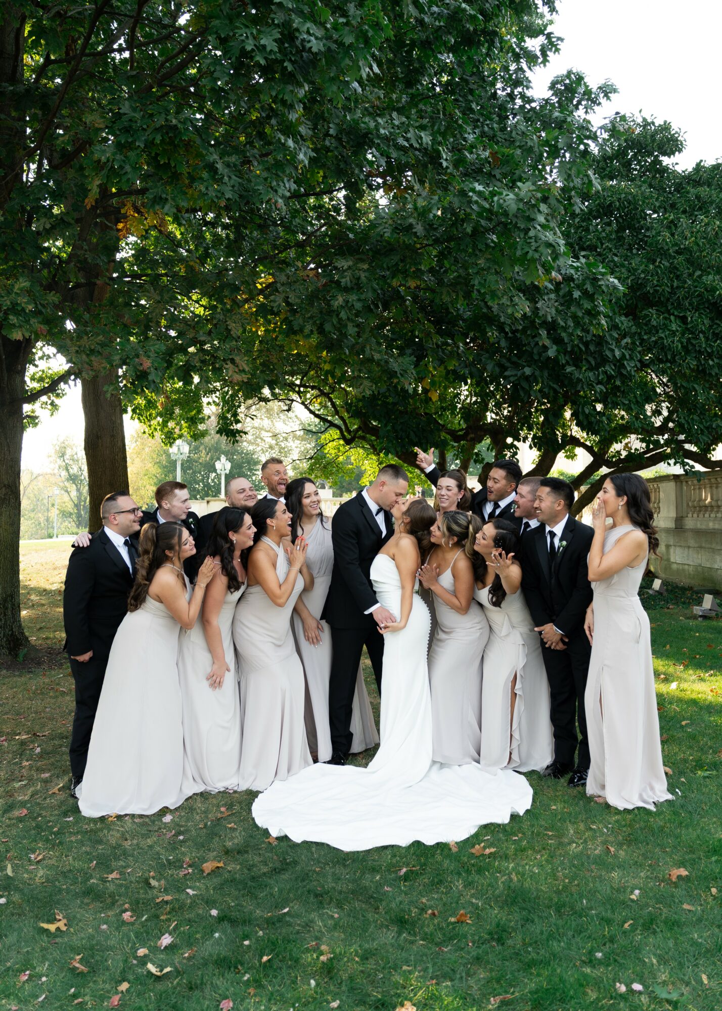 Group of people in formal attire outdoors under a large tree, some holding hands, with a white dress on the ground.