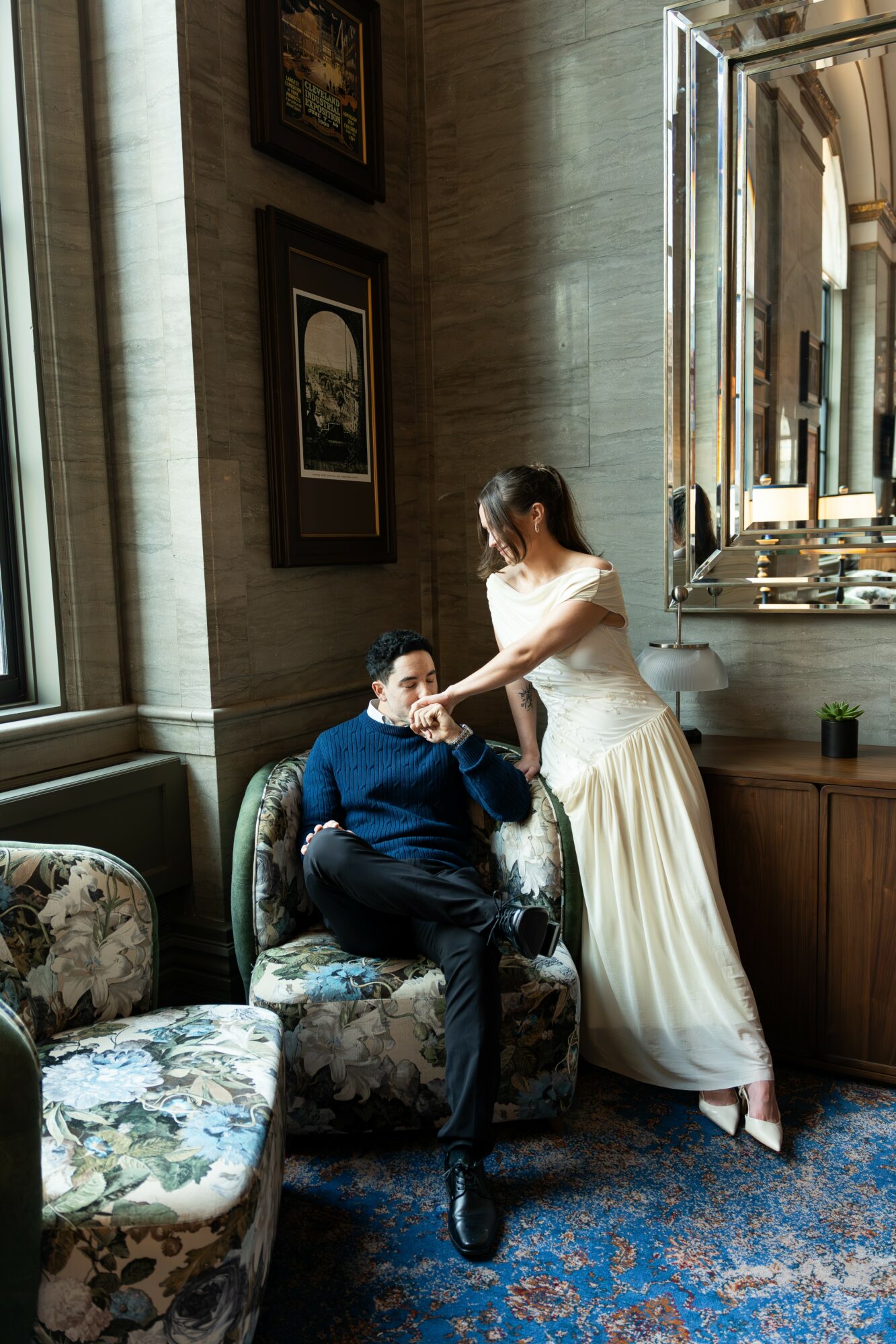 Woman in white dress standing next to seated man in dark clothing, inside a room with large windows and framed pictures.