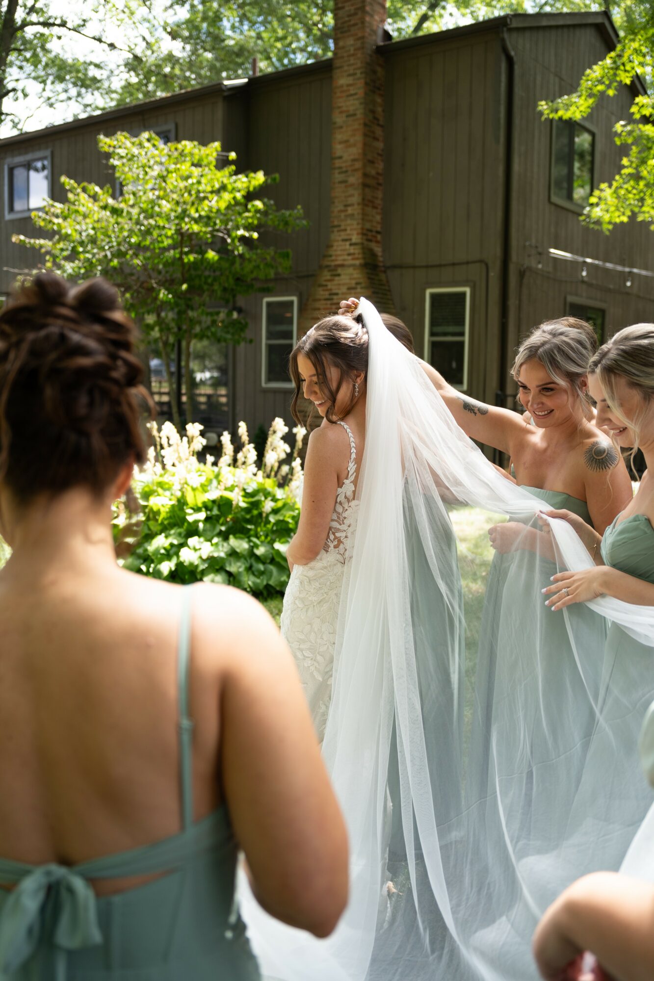 Bride in wedding dress with veil surrounded by women outdoors near a dark green building and trees.
