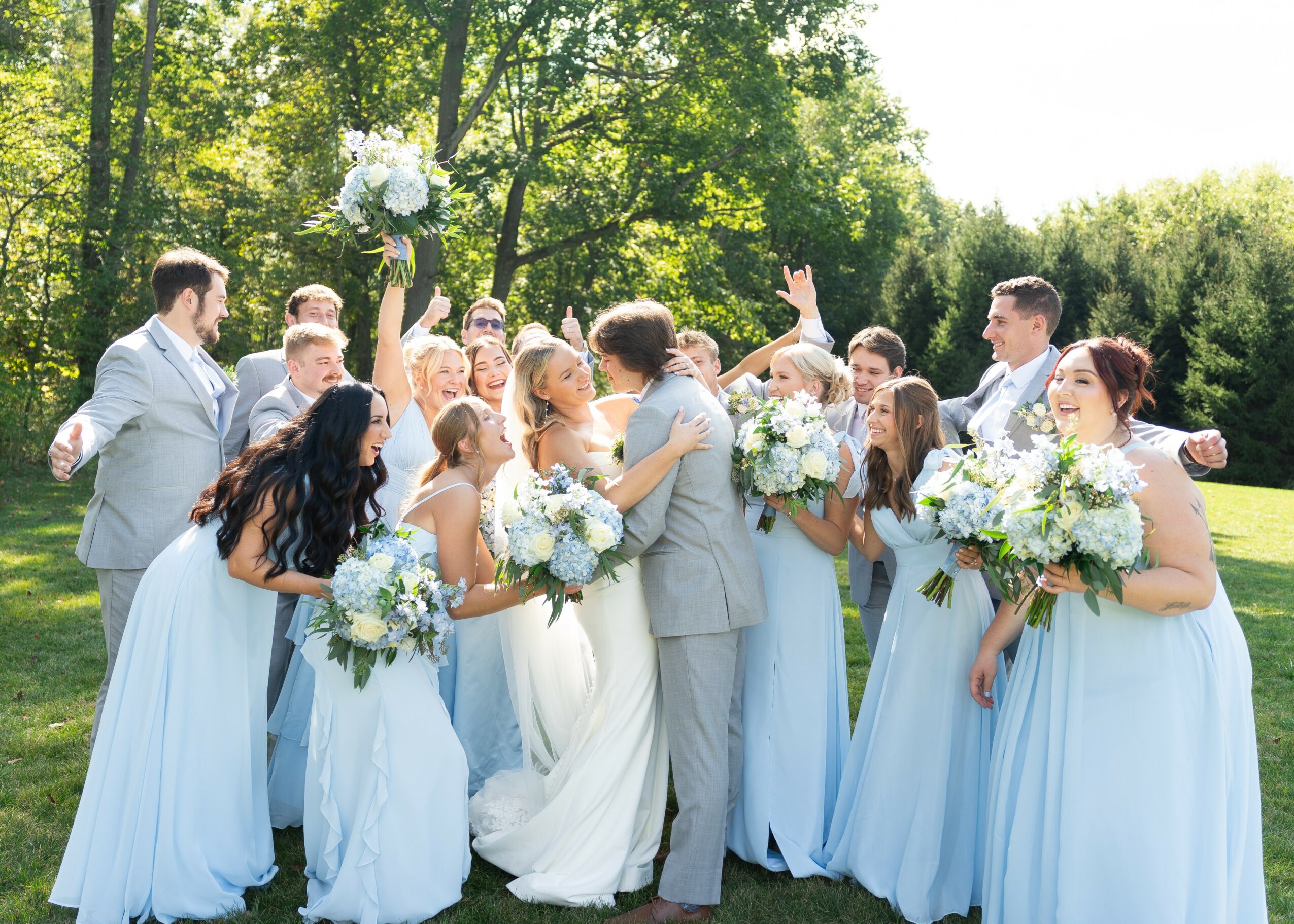 Group of people outdoors, dressed in formal attire, holding bouquets, celebrating a wedding in a green park.
