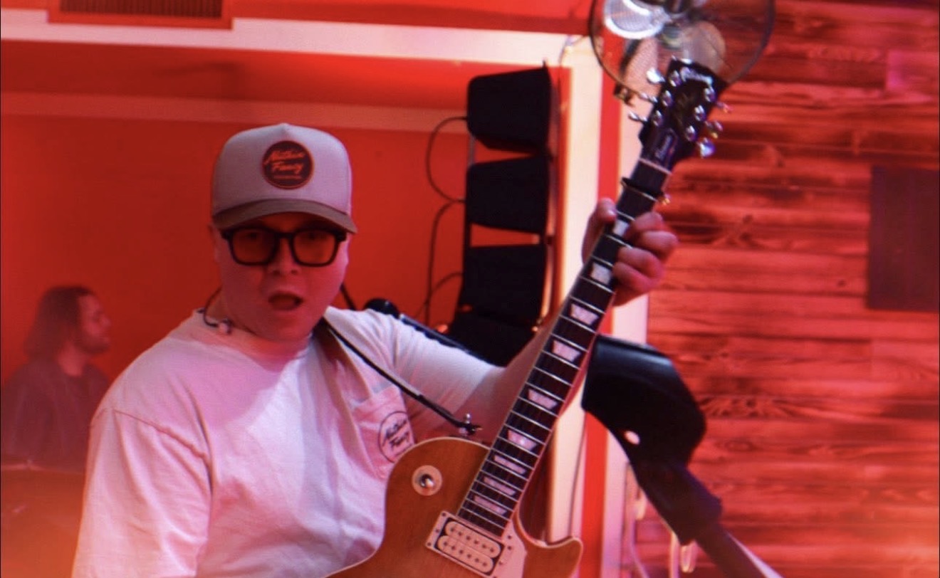 Person wearing glasses, baseball cap, and white shirt holding an electric guitar in a red-lit room.