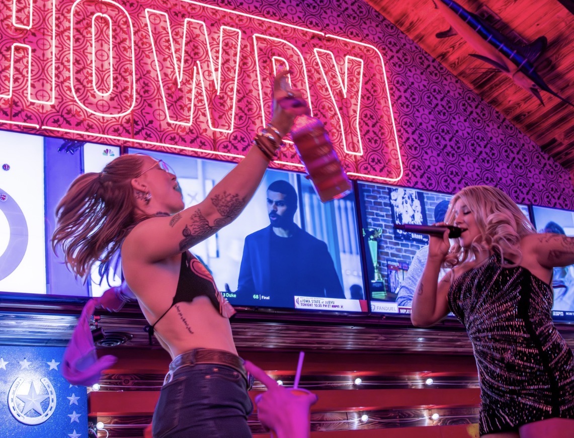 Two women singing and dancing in a karaoke bar with bright neon signs and screens in the background.