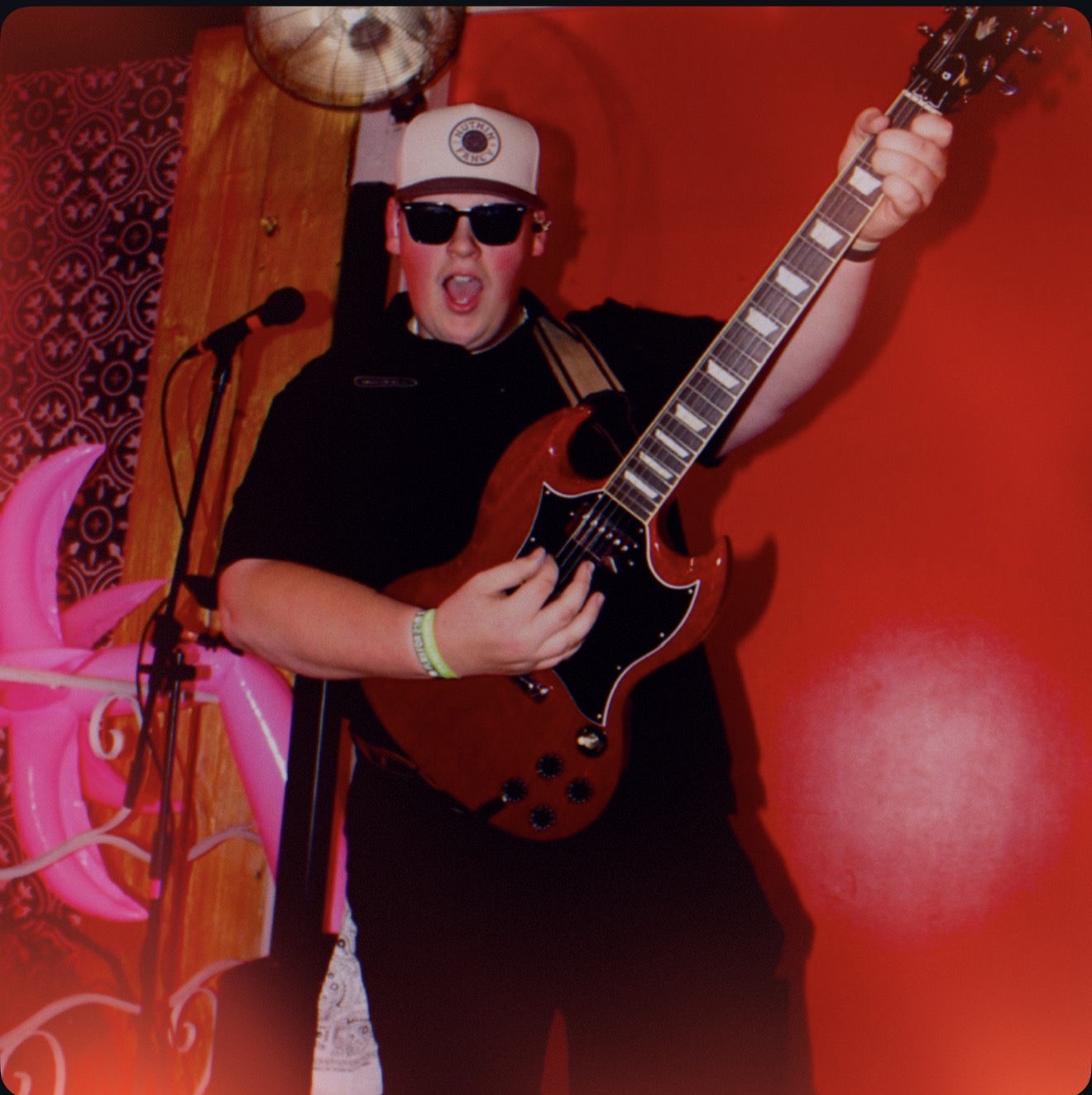 Person wearing sunglasses and a cap, holding an electric guitar, in front of a red background.
