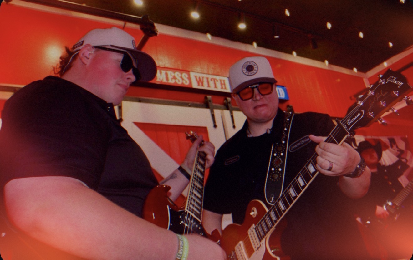 Two musicians playing guitars in a red-lit room, wearing sunglasses and caps, with a sign in the background.