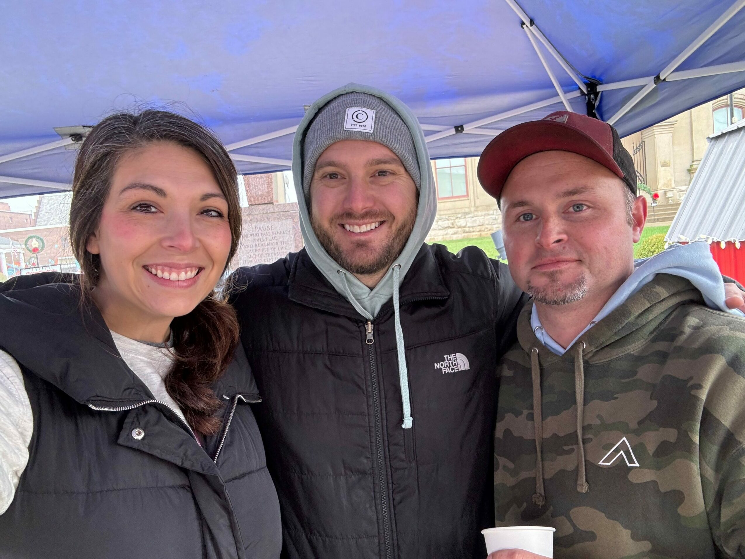 Three people smiling, standing outdoors under a blue canopy, wearing casual jackets and hats.
