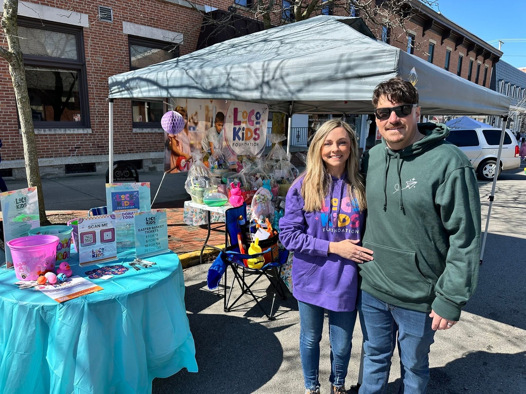 Two people standing outside a booth with a canopy, smiling, with books and items on a table nearby.