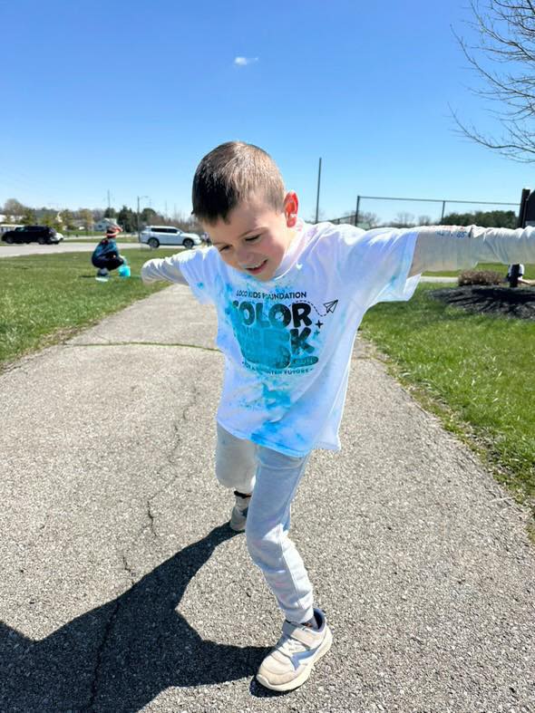 Child running with arms outstretched on a paved path outdoors, smiling, with grass and trees in background.