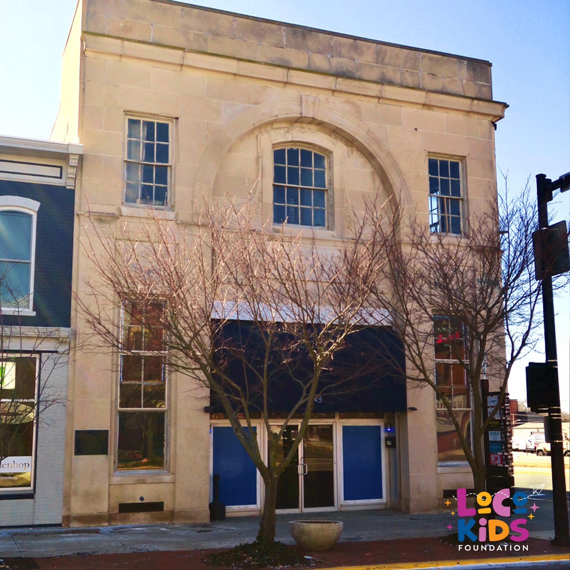 Three-story building with large windows and a black awning, leafless trees in front, and a blue door.