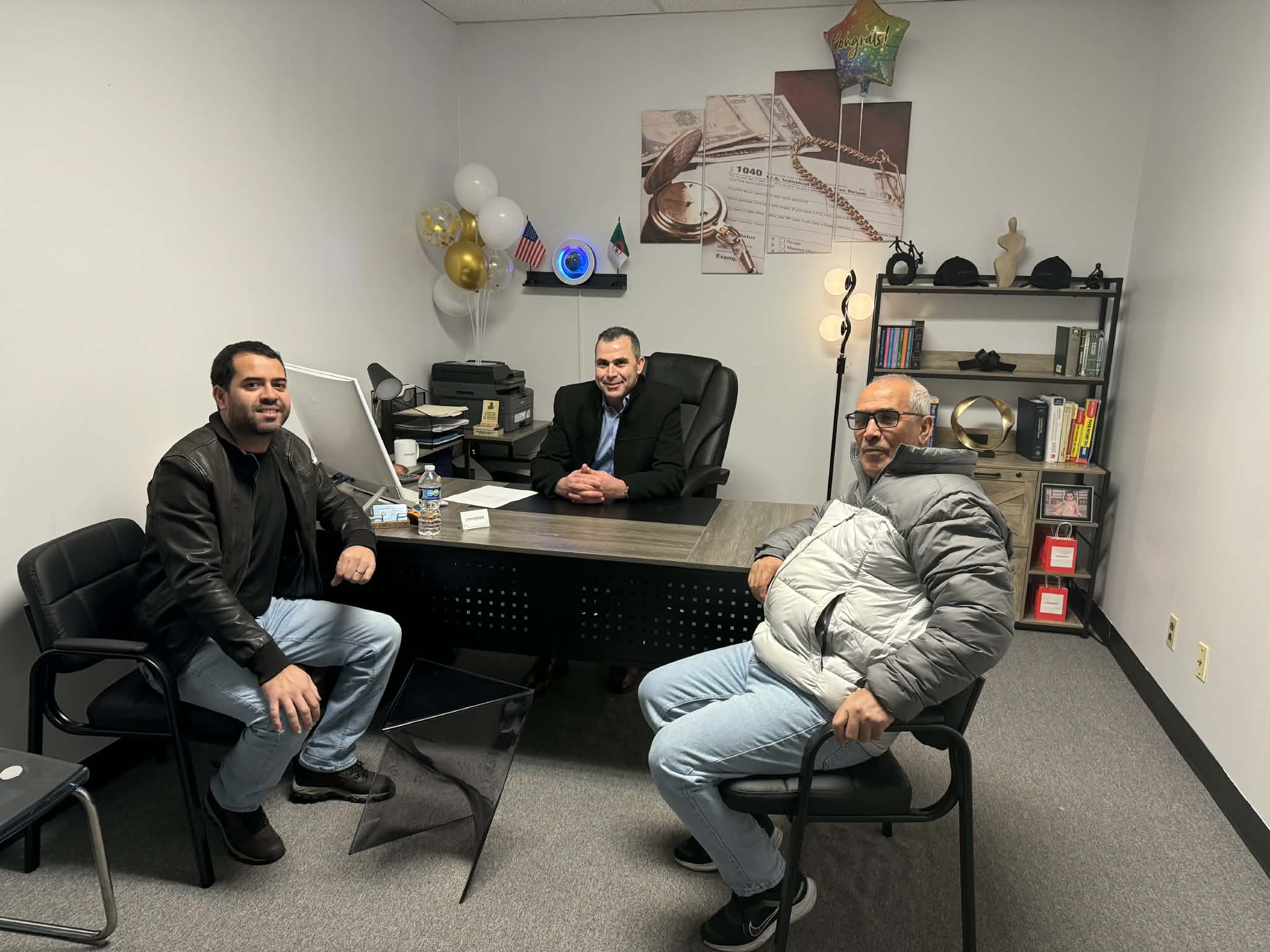 Three men in an office, one seated at a desk and two on chairs, with decorations and shelves in the background.
