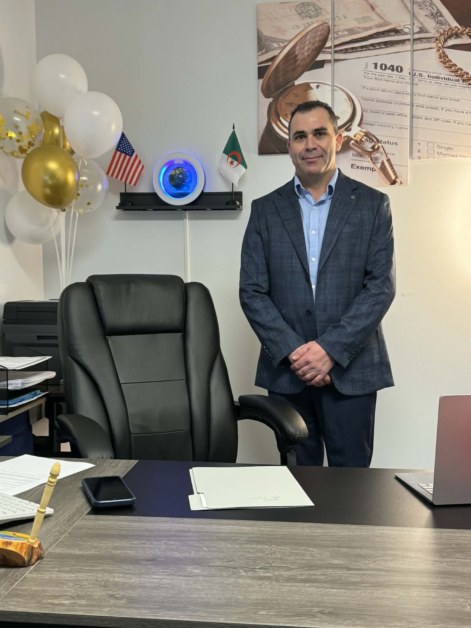 Man in a suit standing in an office with balloons, a laptop, and a large poster of currency notes on the wall.