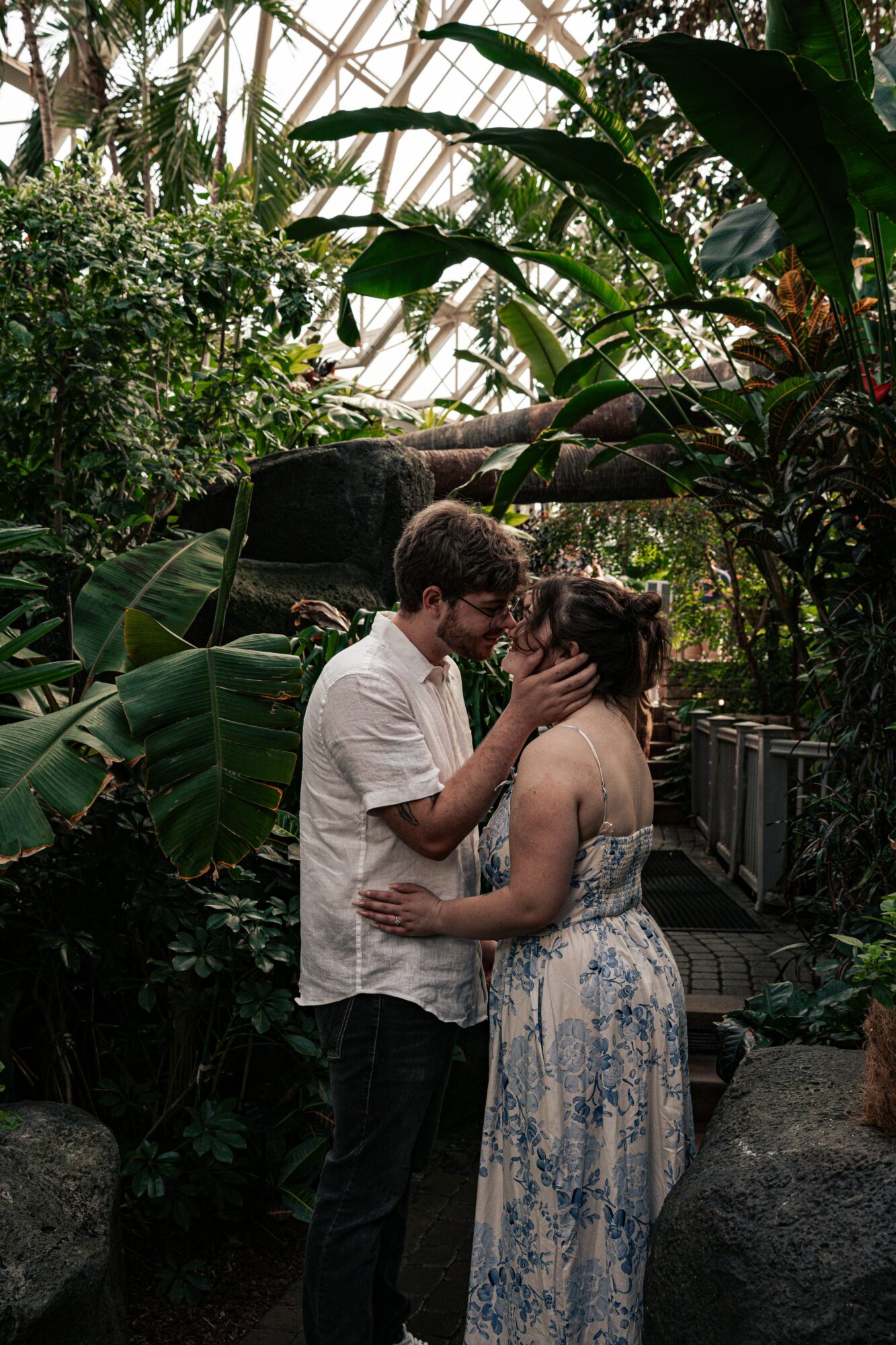 A couple stands close, touching foreheads and holding hands in a lush, green garden setting.