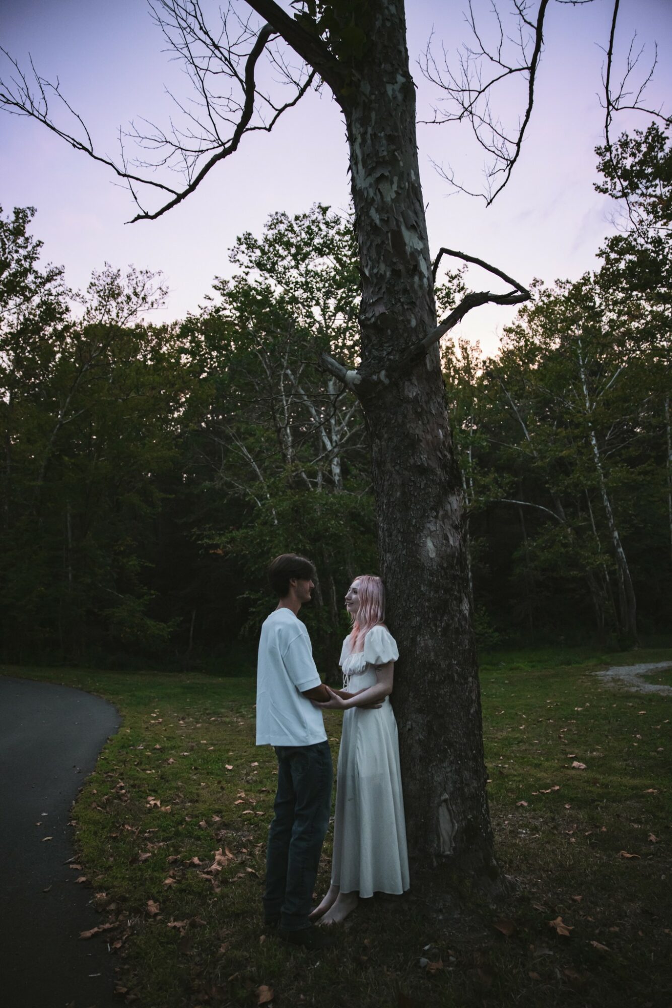 Two people stand near a large tree in a park, surrounded by greenery and a clear sky.