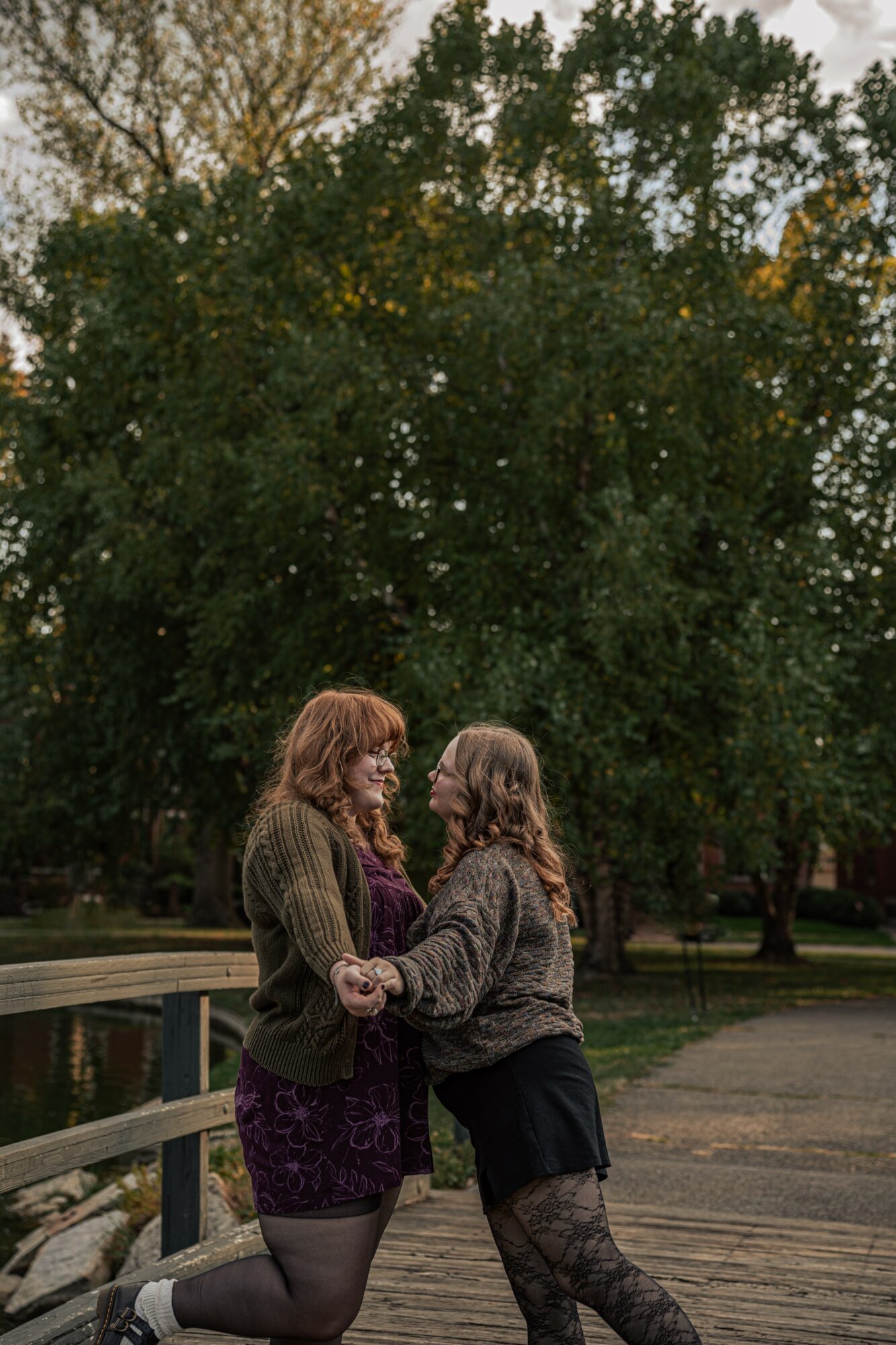 Two people holding hands and facing each other outdoors near trees and a wooden walkway.