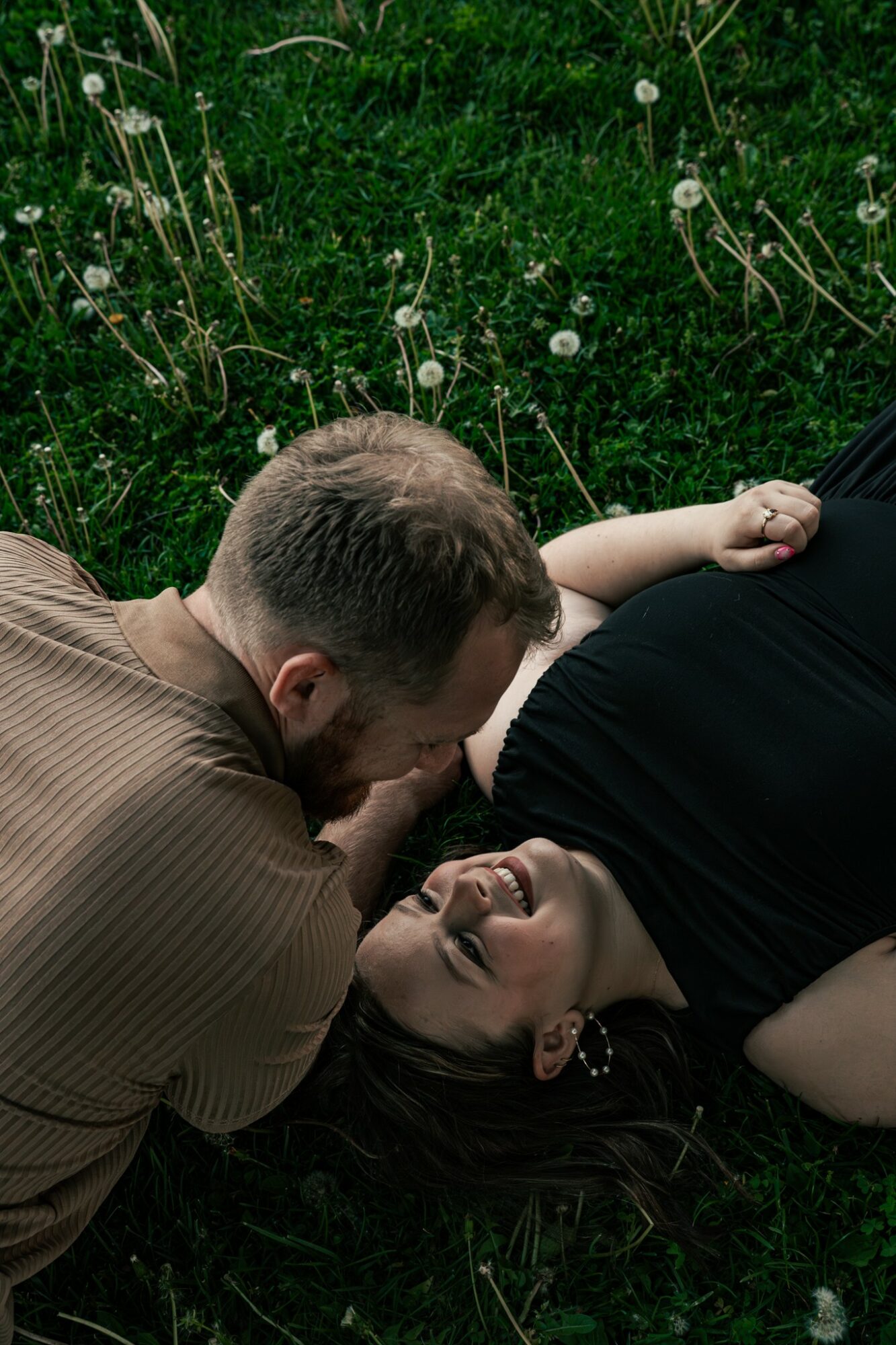 Two people lying on grass, smiling and looking at each other, surrounded by dandelions.