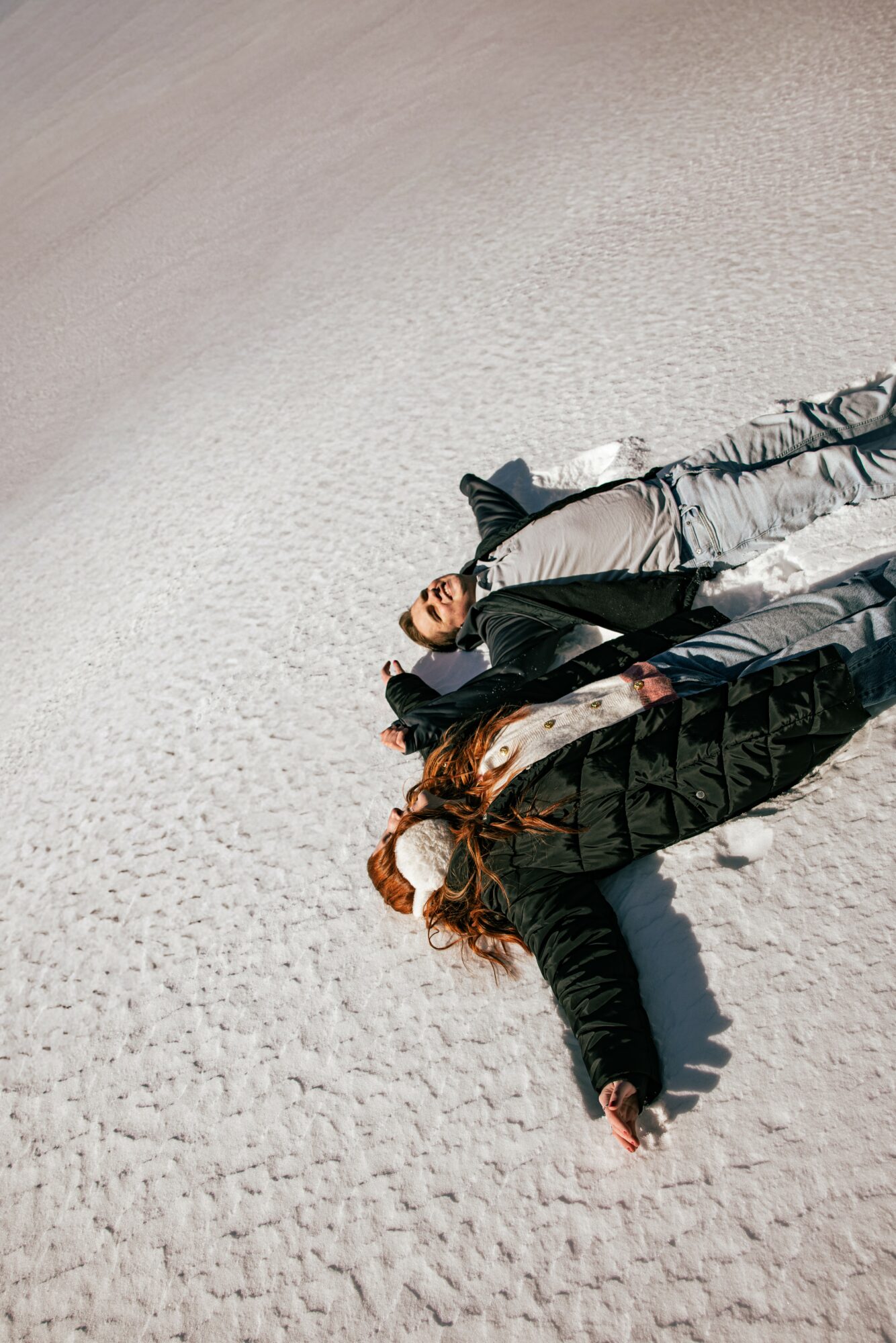 Two people lying on sandy ground, one with long hair and the other with short hair, both wearing dark jackets.