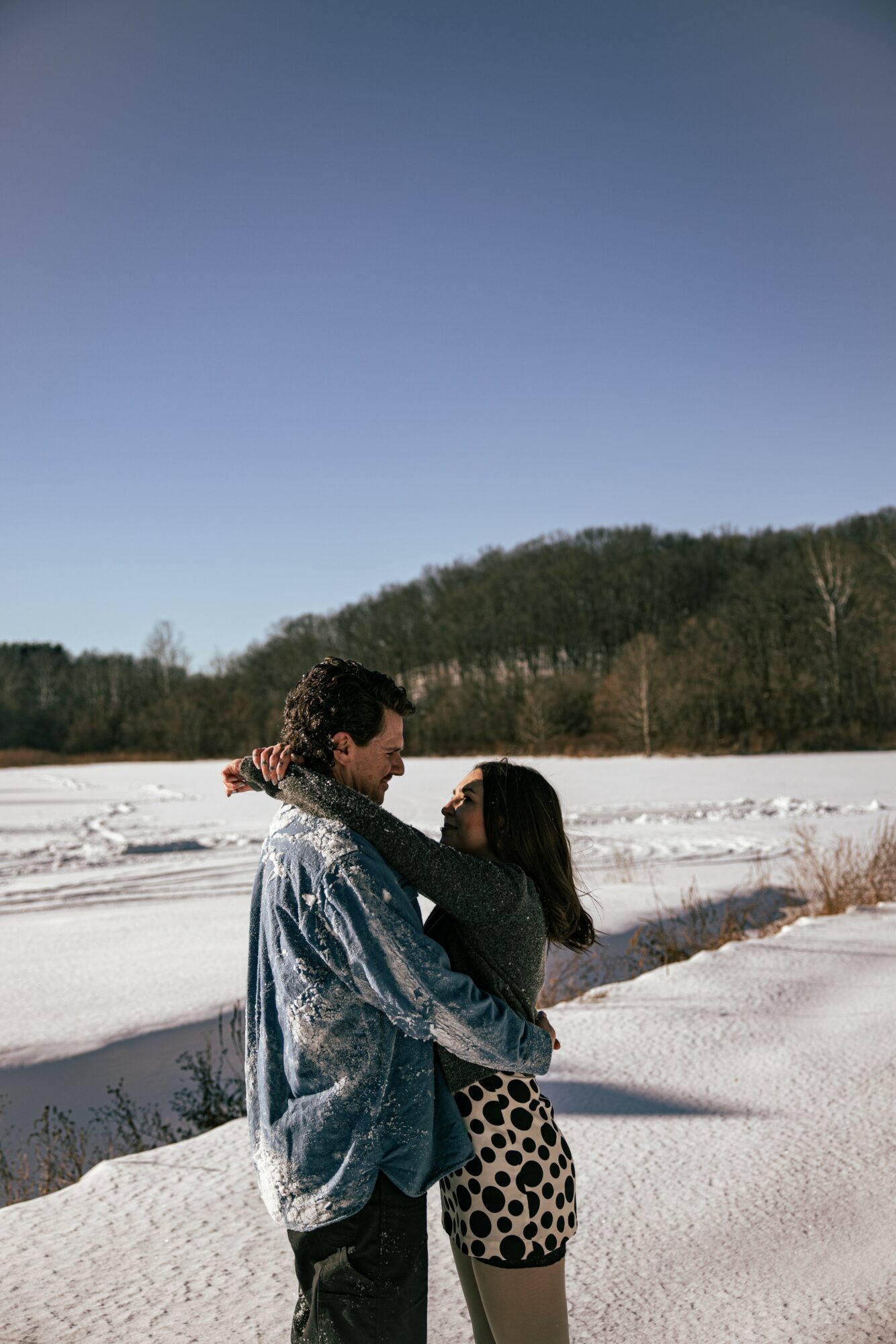 A couple stands close together in a snowy landscape with a forested hill and clear blue sky behind them.