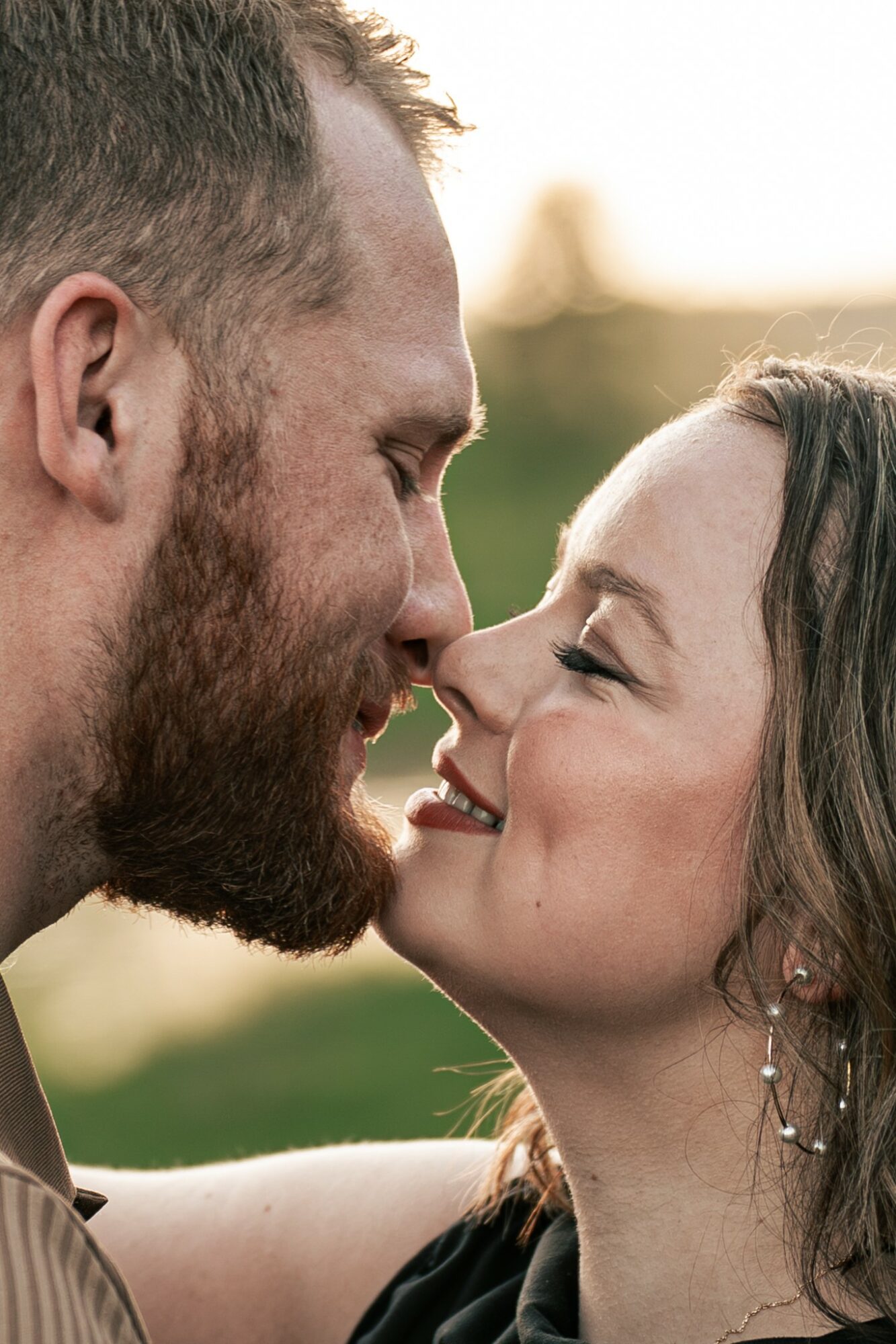A man and woman face close, smiling, with noses touching outdoors during sunset.
