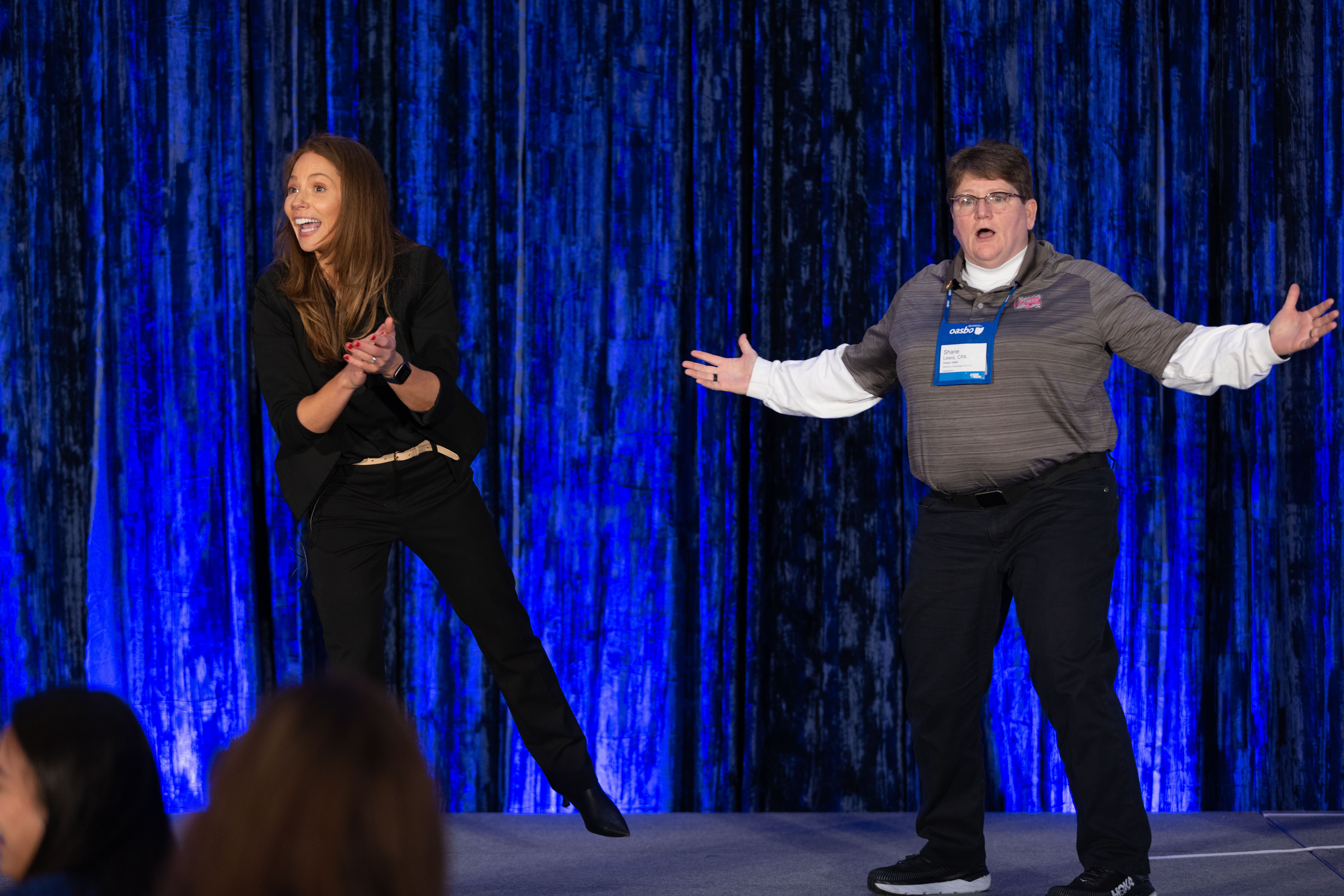 Two people on stage with a blue curtain background, one clapping and the other gesturing with arms outstretched.