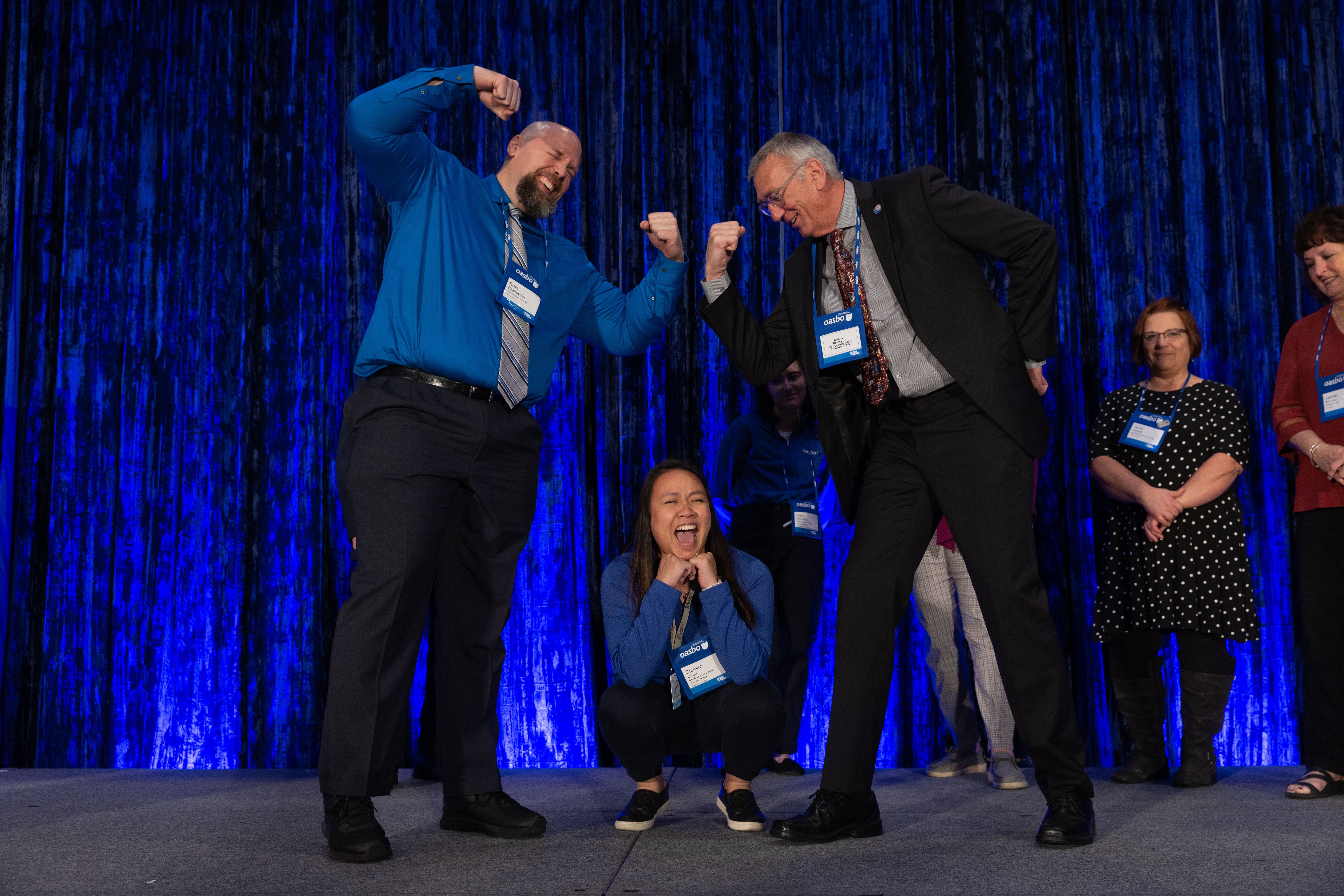 Two men on stage flex their arms in a friendly competition, with three women standing behind them, smiling.