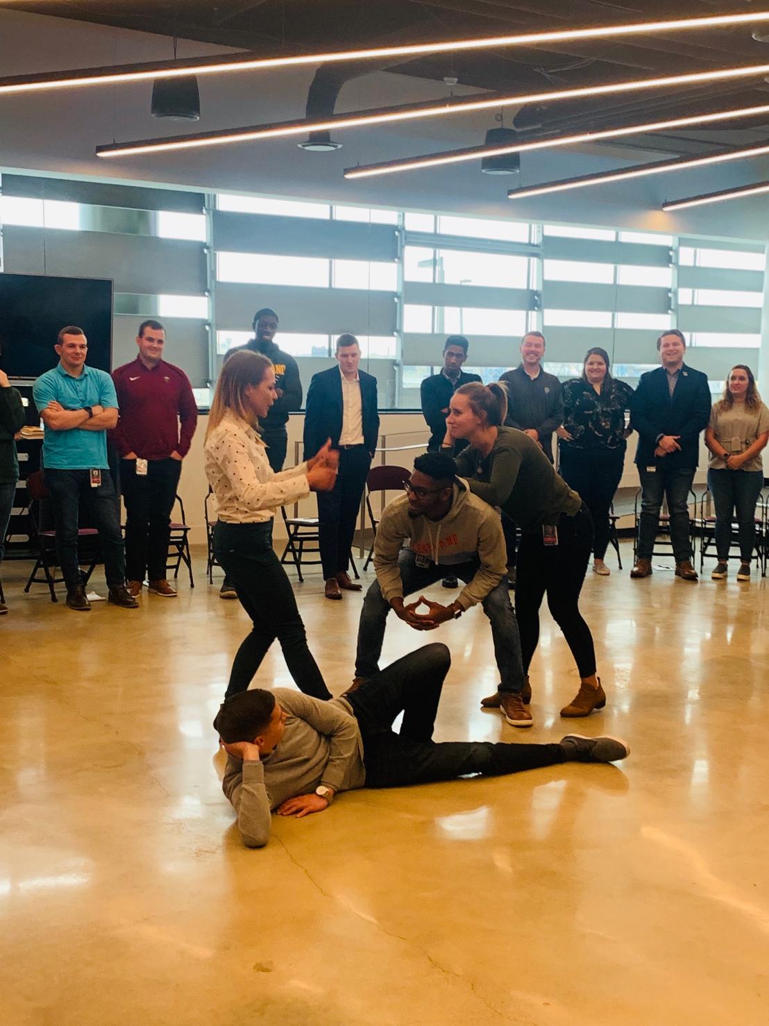 Group of people in a spacious room with large windows, some standing and others on the floor, engaging in an activity.