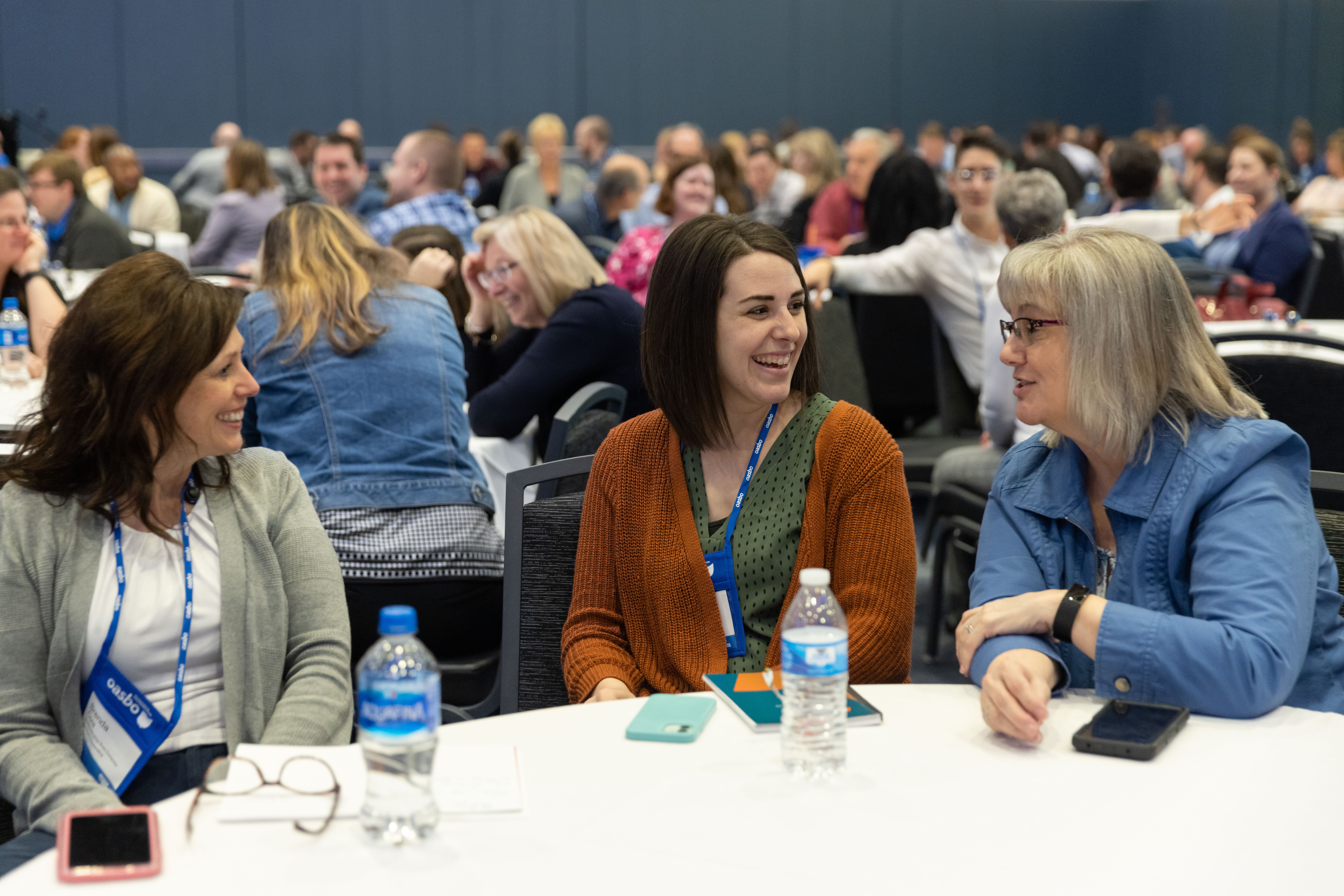 Three women sitting at a table in a large conference room, engaged in conversation, with many people in the background.