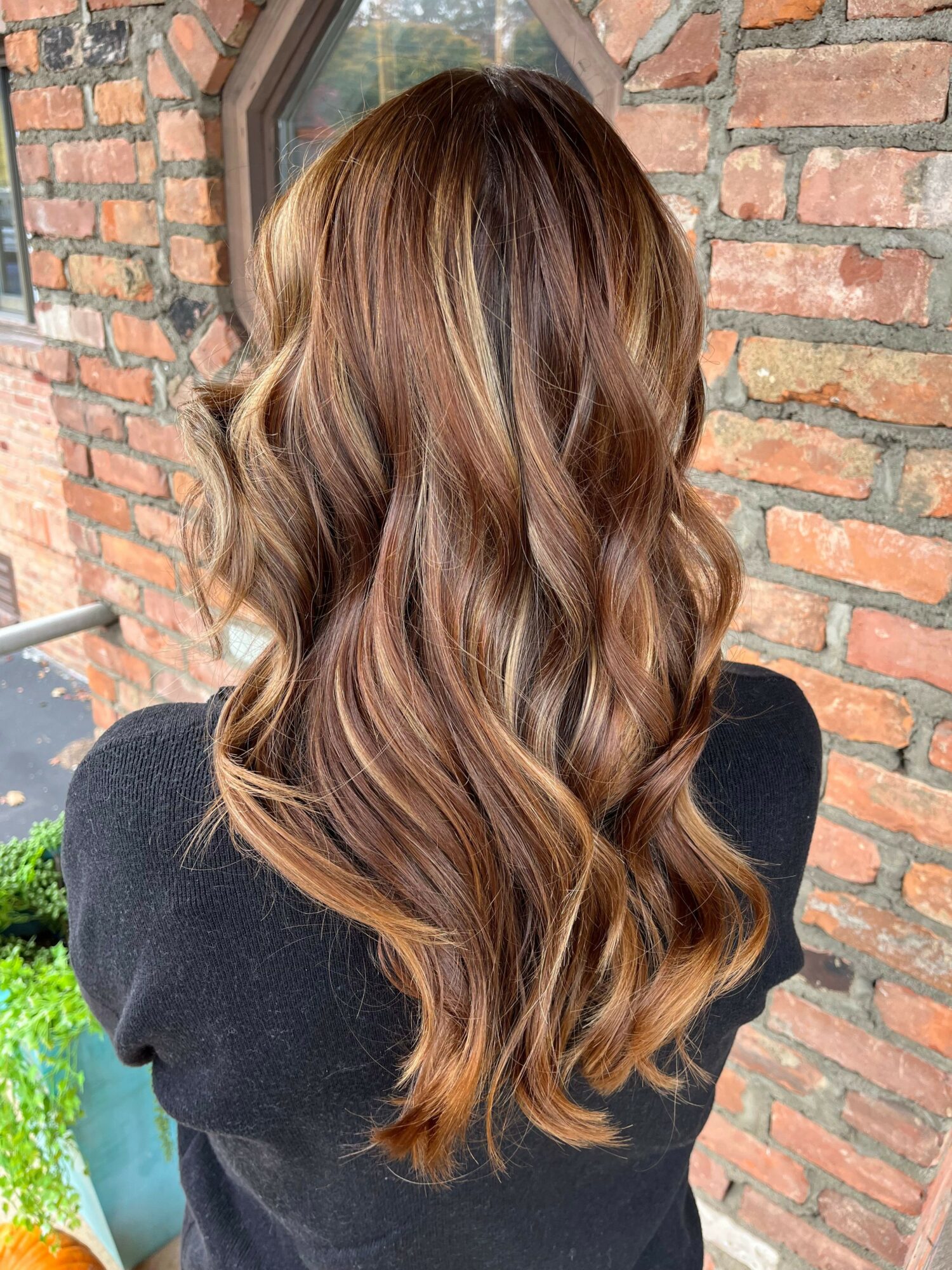 Back of a woman with long, wavy, brown hair with highlights, standing outdoors near a brick wall.