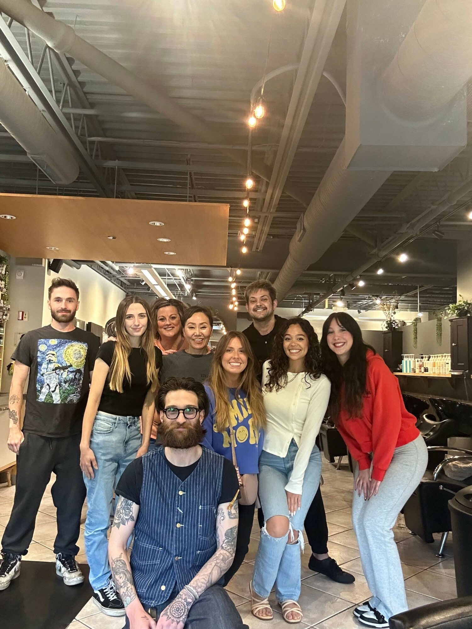 Group of eight diverse young adults standing together indoors, smiling, with industrial ceiling and lighting above.