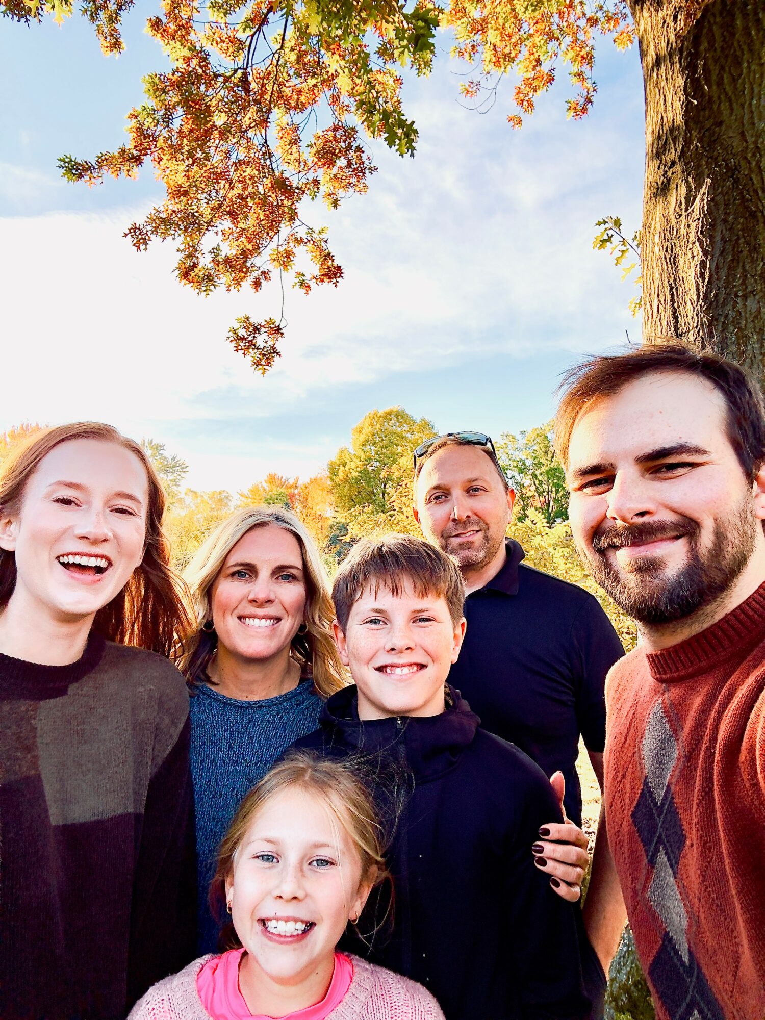 Group of six smiling people outdoors under a tree with autumn leaves, sunny sky in background.