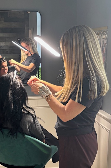 A hairstylist with blonde hair works on a client's hair in a salon, with a mirror reflecting the scene.