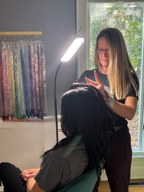 Two women, one with long blonde hair and glasses, the other with dark hair, in a room with a window and fabric samples.