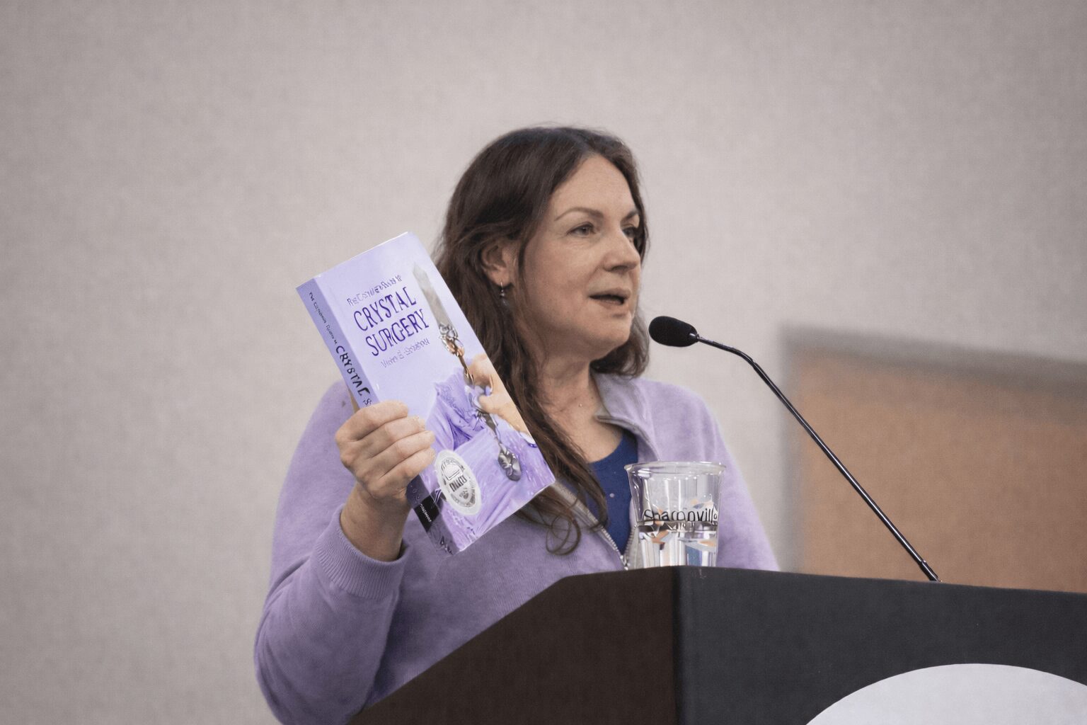 Woman speaking at a podium, holding a brochure, with a glass of water nearby, against a plain background.