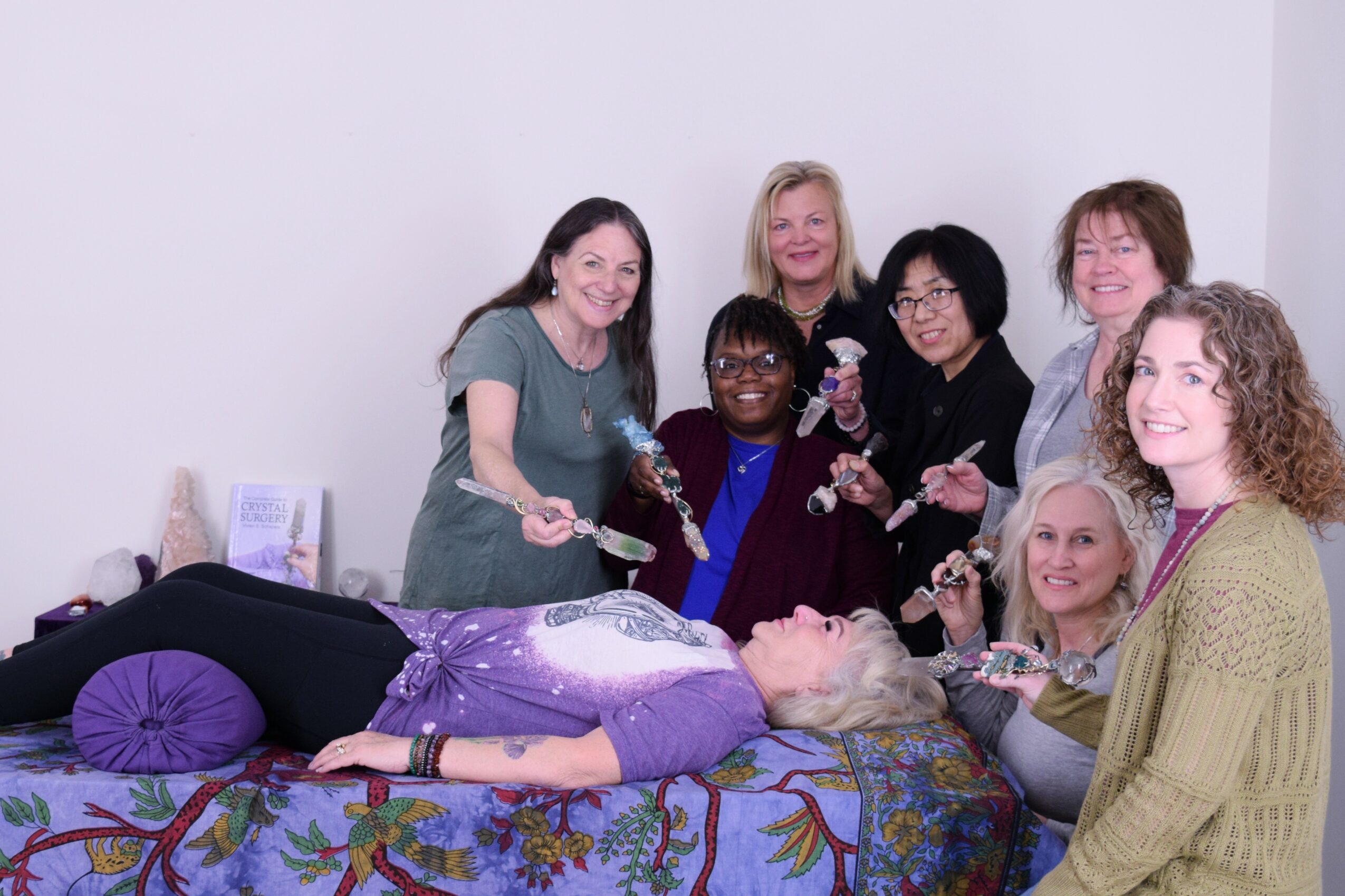 Group of seven women gathered around a person lying on a bed, smiling and holding glasses, in a bright room.