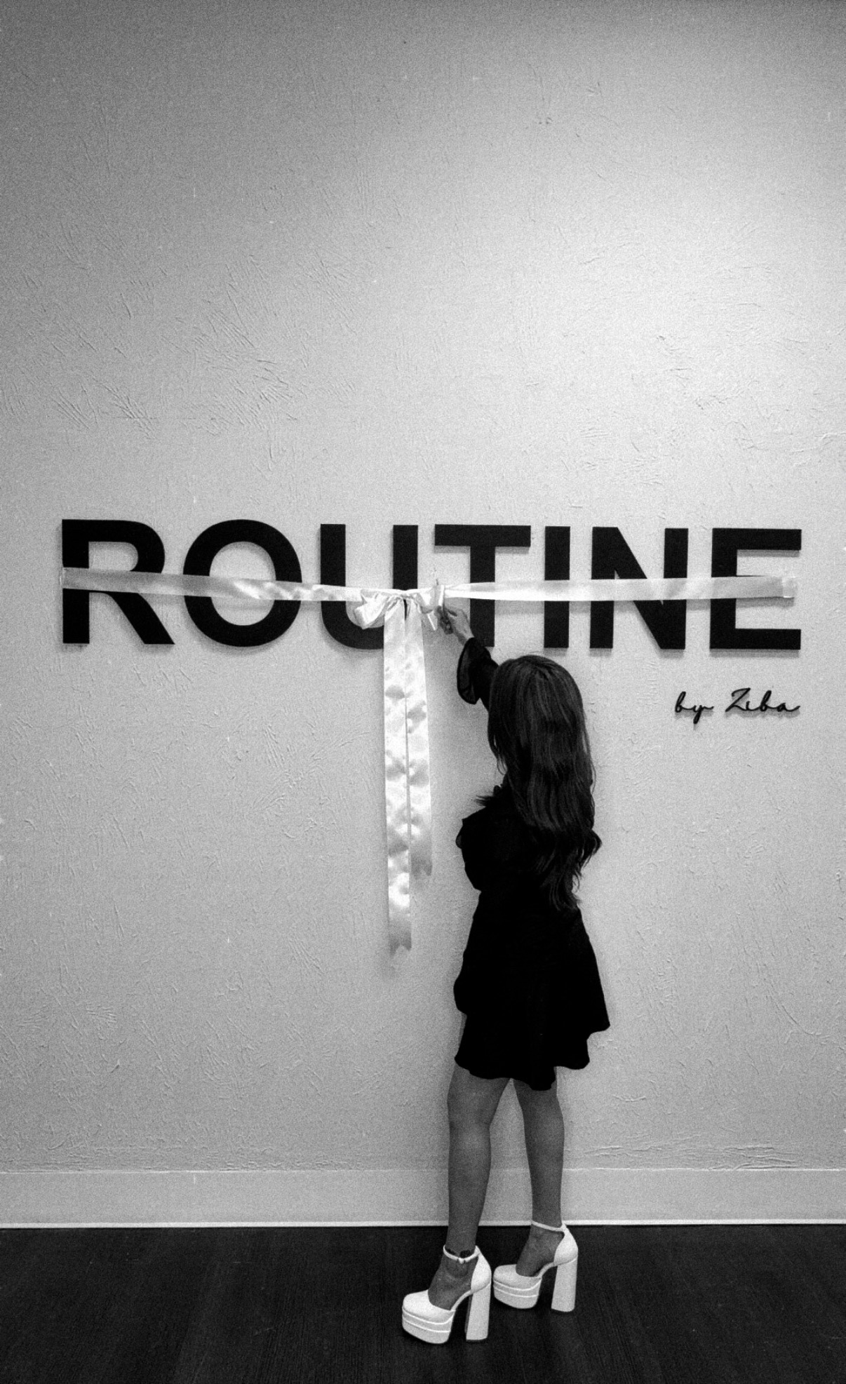 A girl in a black dress and high heels stands in front of a wall with the word 'ROUTINE' and a signature.