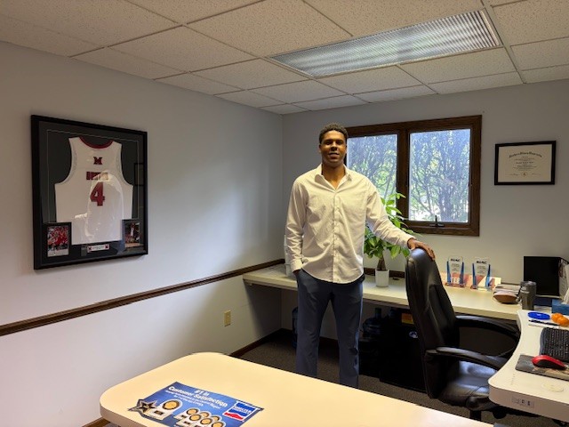 Man standing in an office next to a desk with a window behind him, framed sports jersey on wall, and certificate on wall.