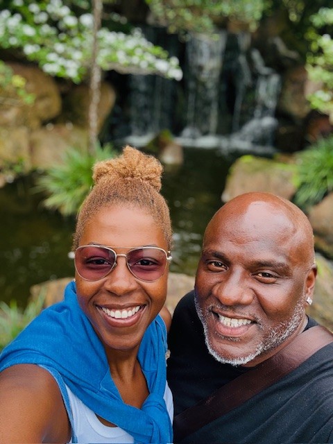 Two smiling people taking a selfie outdoors with a waterfall and greenery in the background.