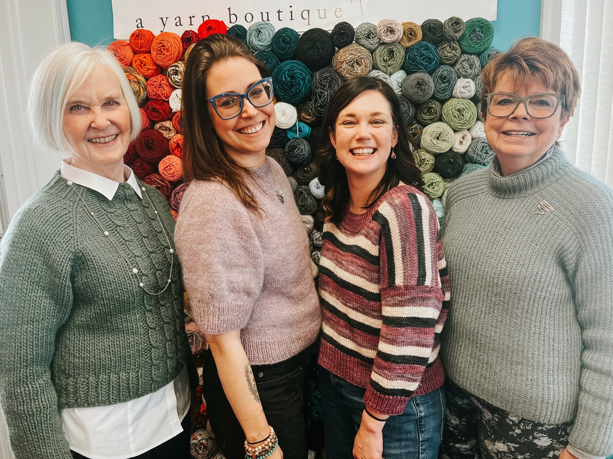 Four women standing in front of a colorful yarn display, smiling at the camera.