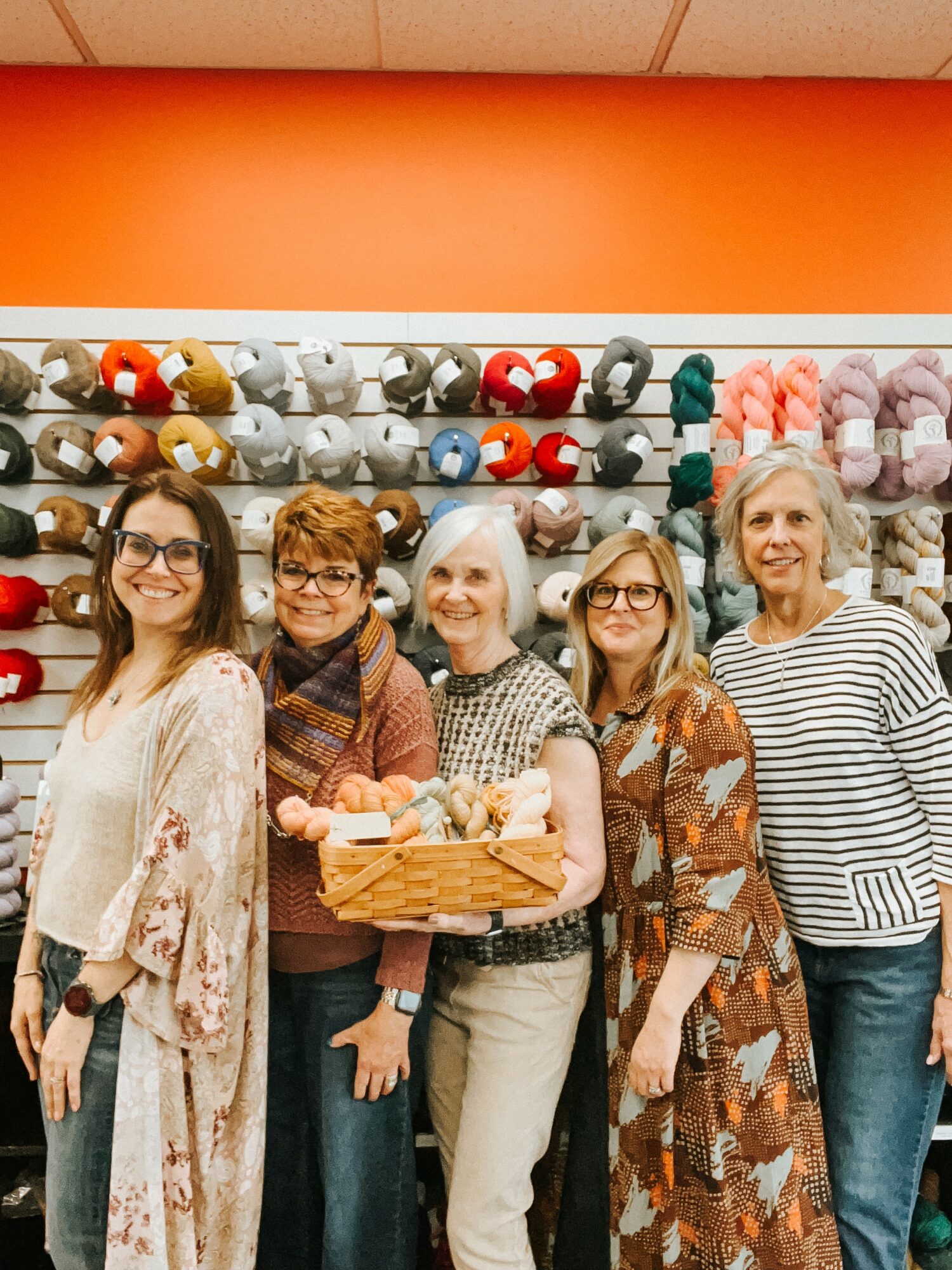 Five women standing in front of shelves with yarn and knitting supplies, smiling, in a craft store.