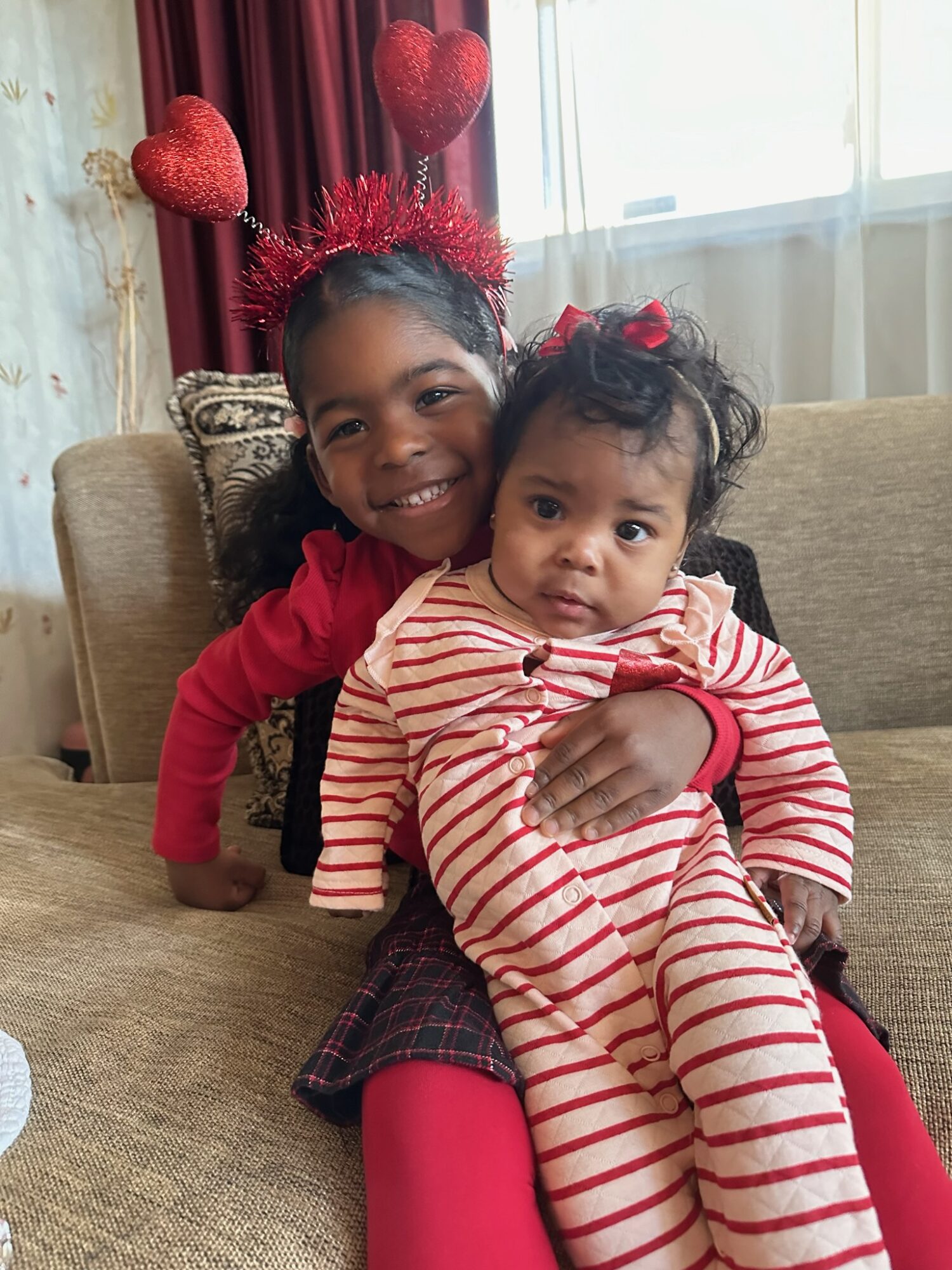 Two young girls sitting on a sofa, one smiling and wearing a red headband with heart-shaped decorations, the other in striped pajamas.